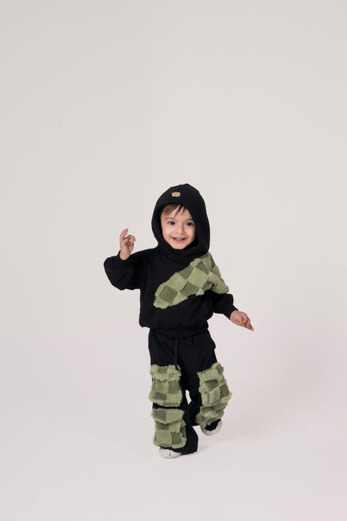 Smiling toddler in black hoodie and pants with green checkered plush patches on white background