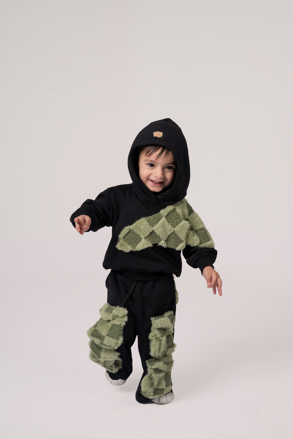 Toddler in stylish black hoodie and pants with textured green checkered patches on plain background