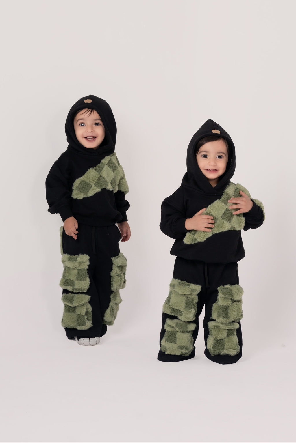 Two toddlers wearing black hooded outfits with green checkered plush patches on top and pants in a minimalist studio