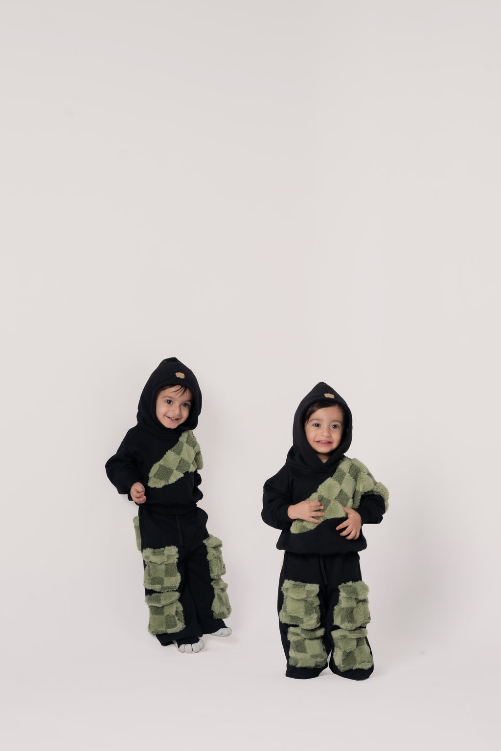 Two toddlers in black hoodies and pants with green textured checkerboard patches on a clean white backdrop