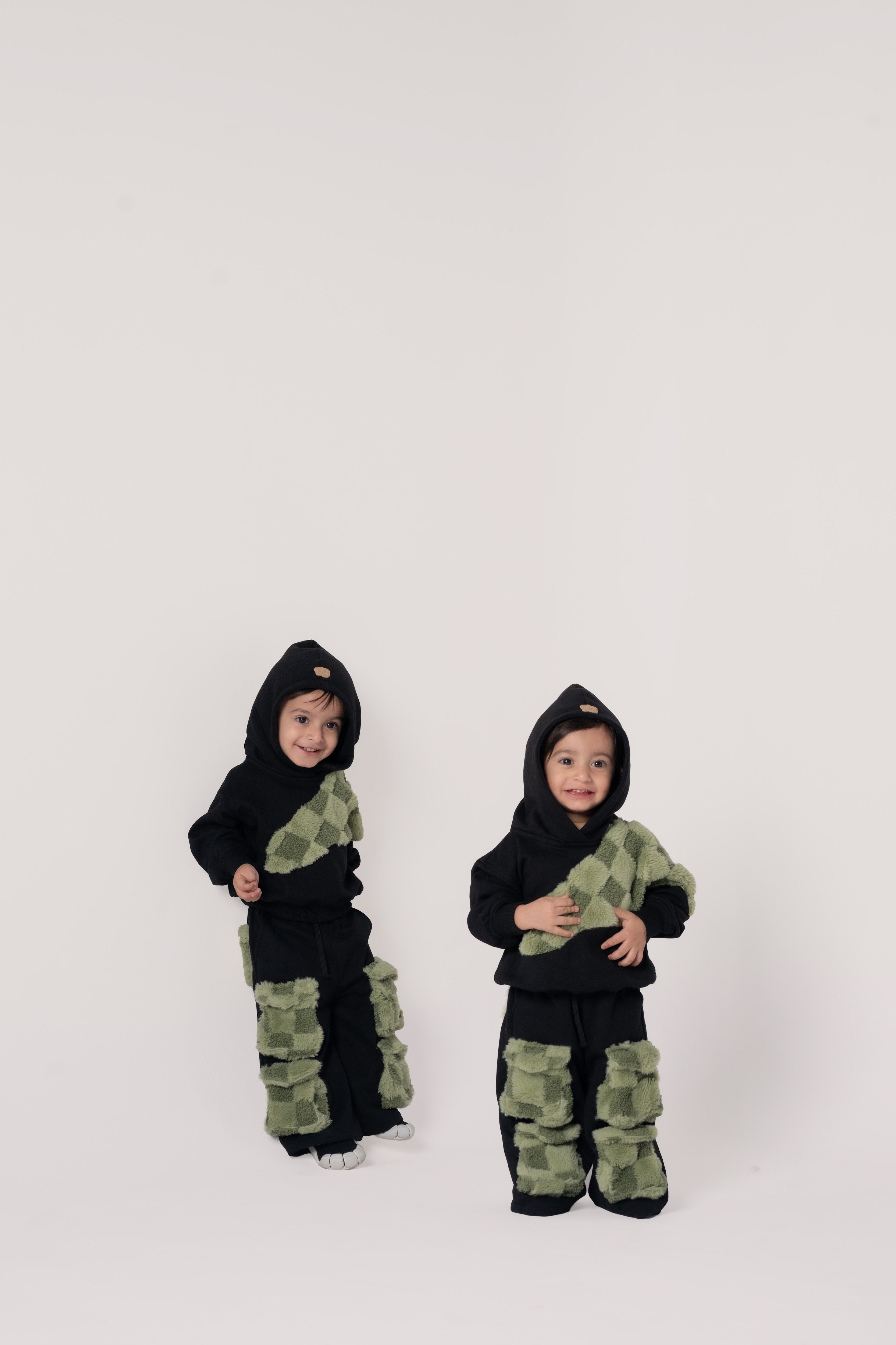 Two toddlers in black hoodies and pants with green textured checkerboard patches on a clean white backdrop