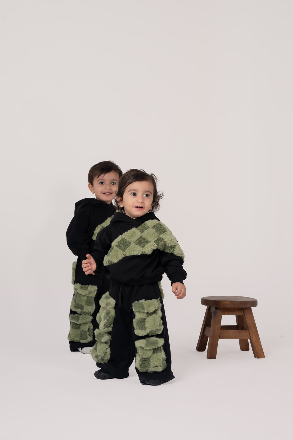 Two toddlers in black outfits with luxurious green checkered fuzzy accents standing near a wooden stool