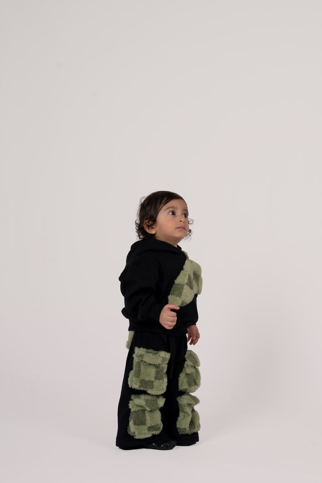 Toddler wearing black outfit with soft green textured patch pockets on pants and hoodie against neutral background