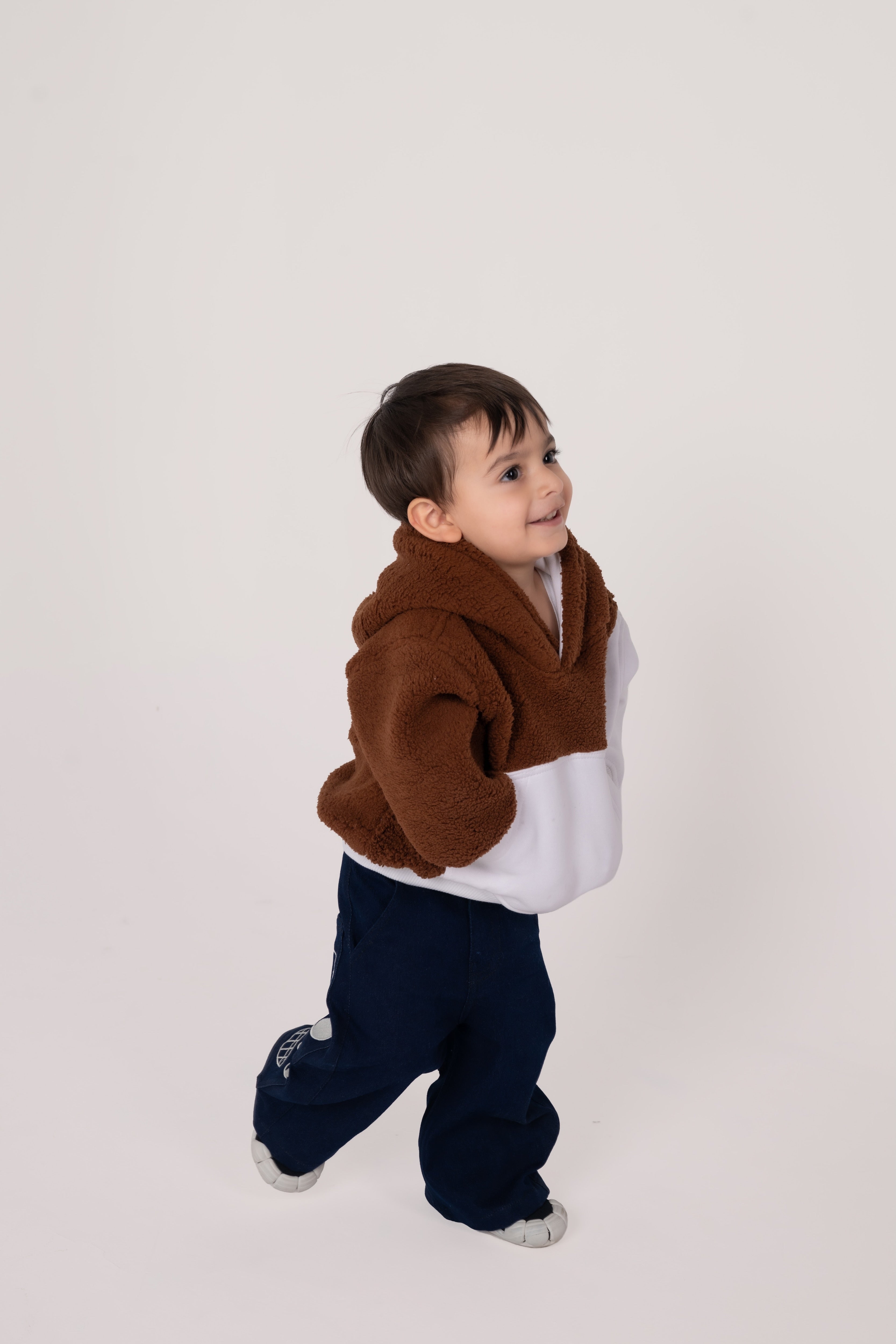 Smiling toddler wearing plush two-tone brown and white hoodie and navy pants on white background