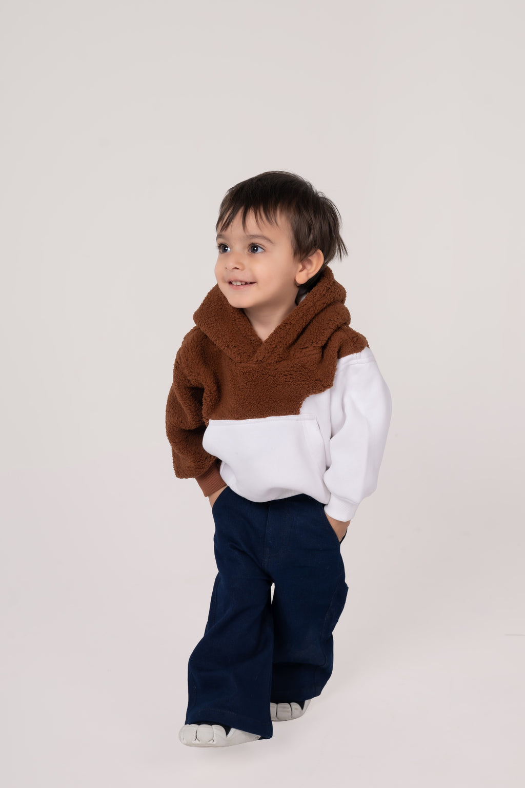 Stylish toddler boy in cozy brown and white hoodie with navy pants on neutral background