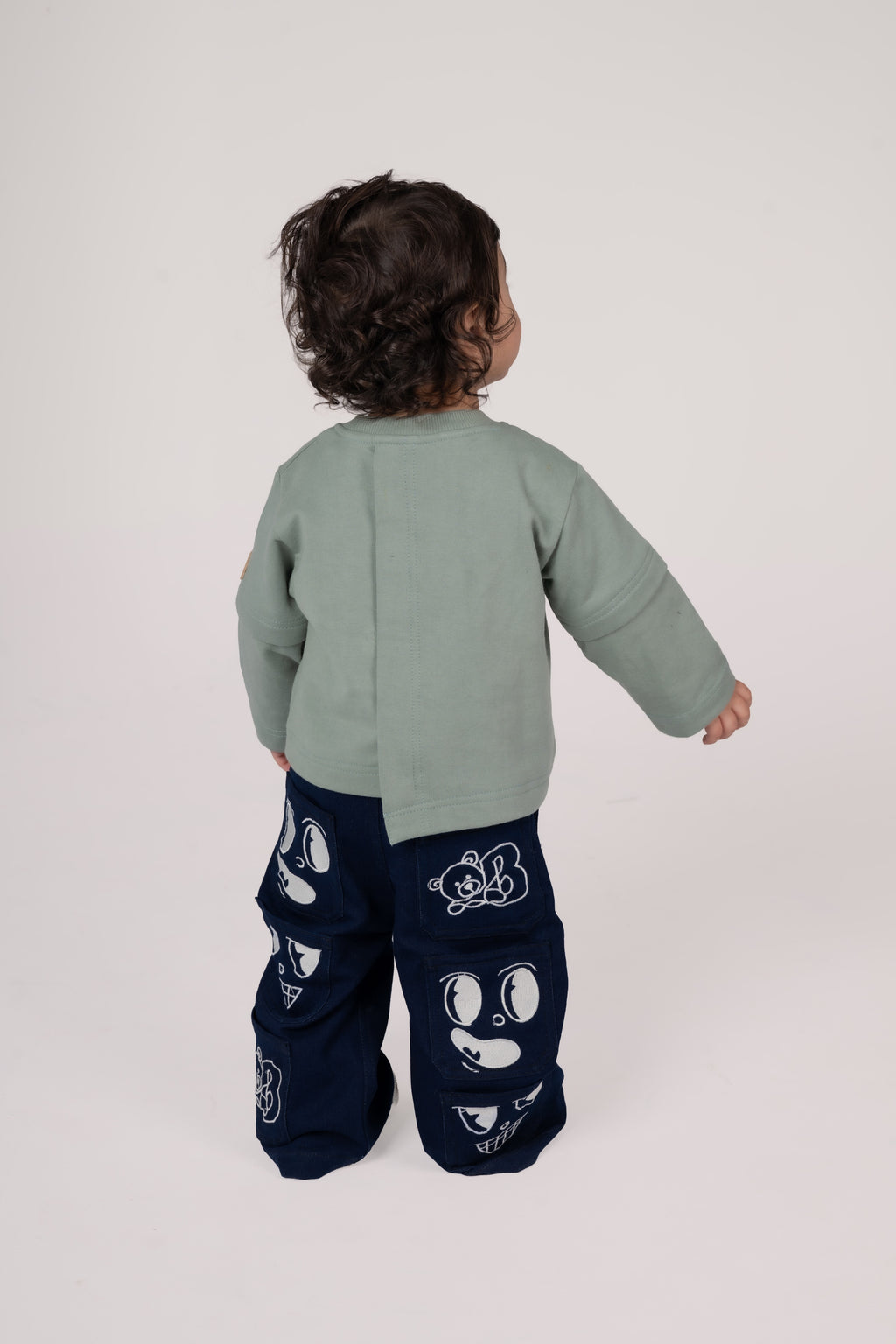 Toddler in sage green sweater and navy pants with playful white cartoon patches on neutral backdrop