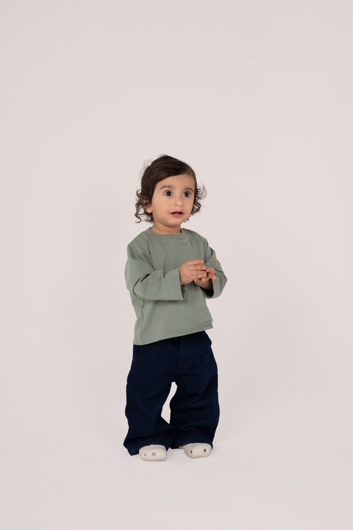 Toddler in sage green top and navy pants with white shoes on soft pink backdrop