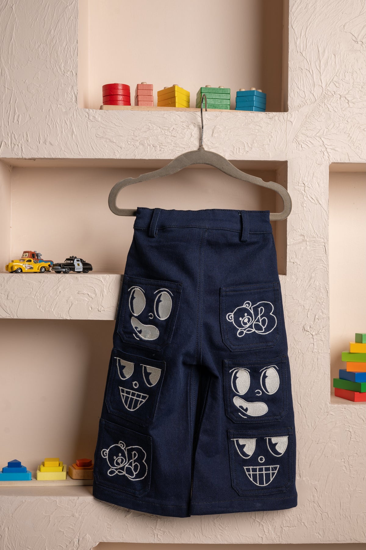 Luxury navy blue kids pants with playful embroidered bear and face designs displayed on a textured beige wall