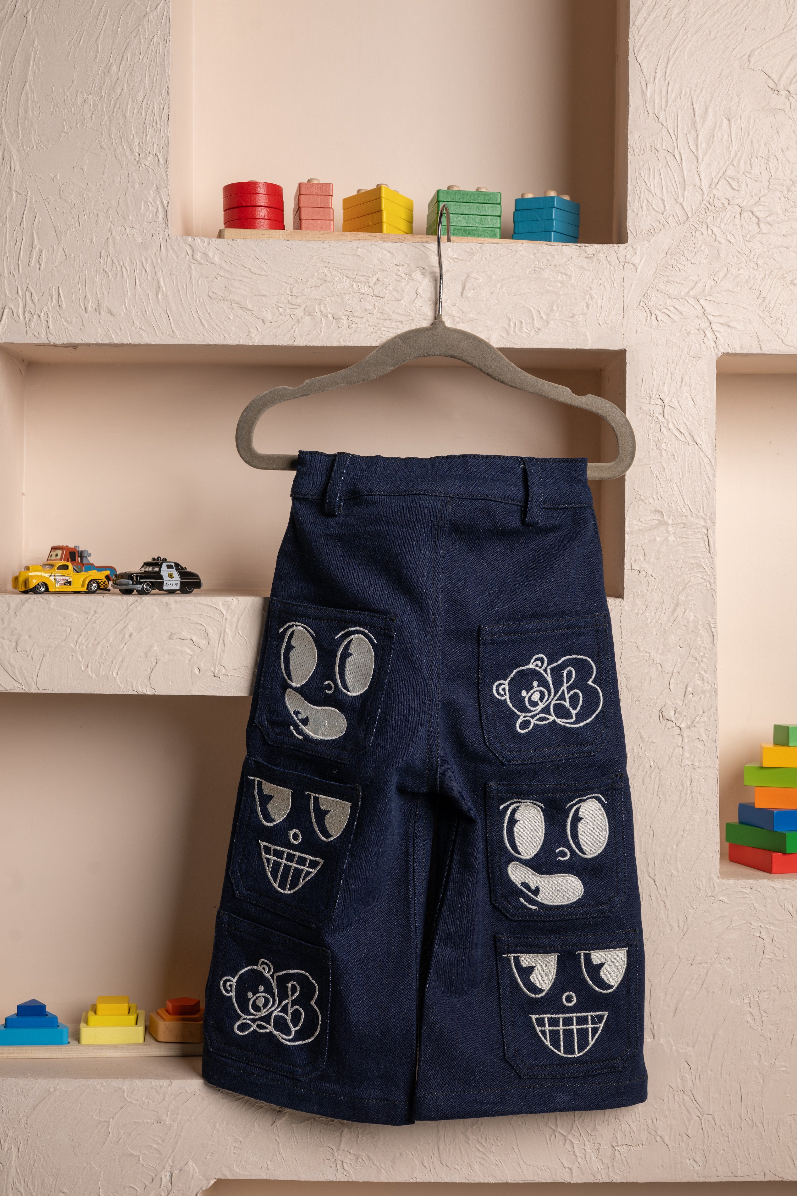 Luxury navy blue kids pants with playful embroidered bear and face designs displayed on a textured beige wall