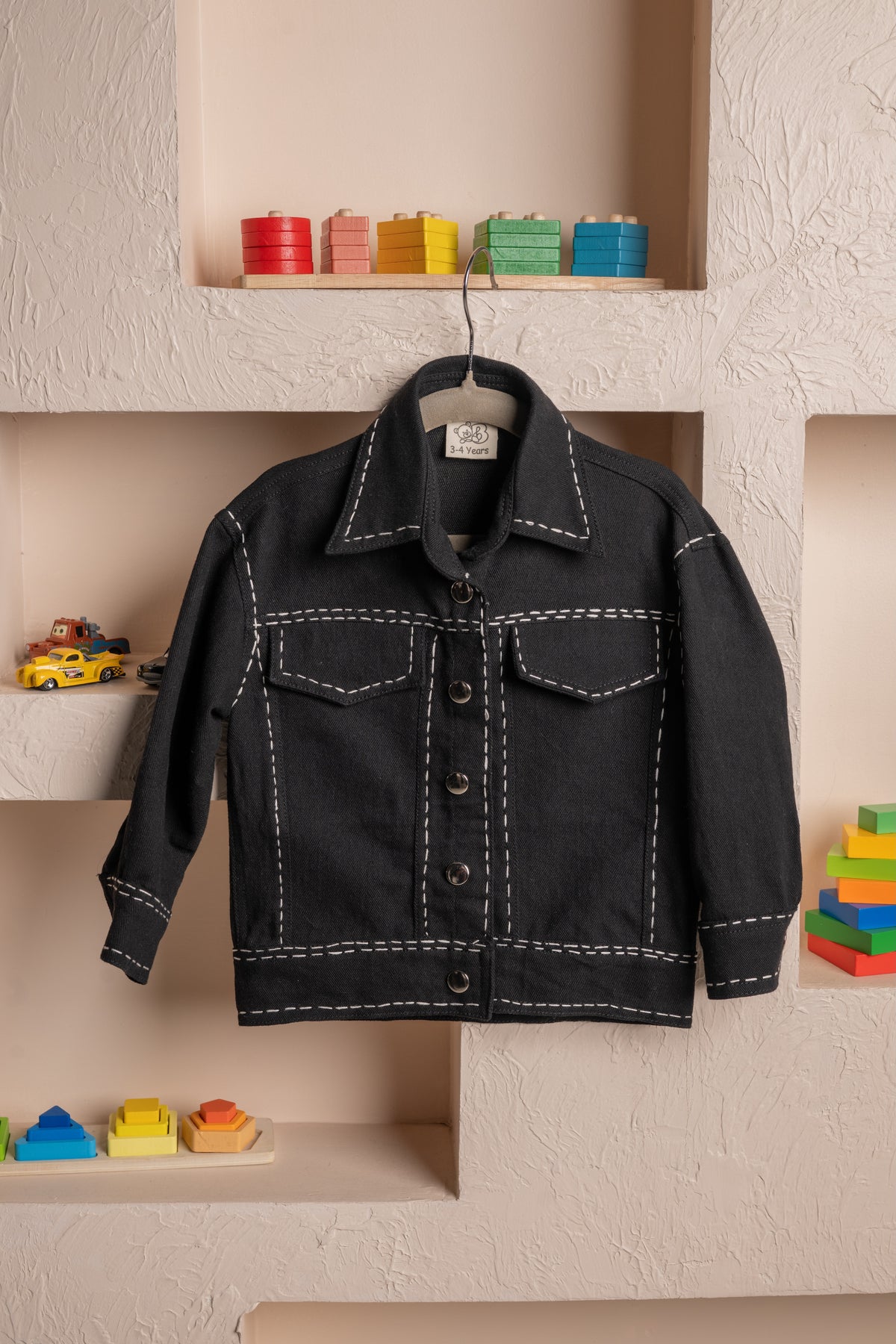 Luxurious black toddler jacket with white stitching on textured beige wall with colorful wooden toys