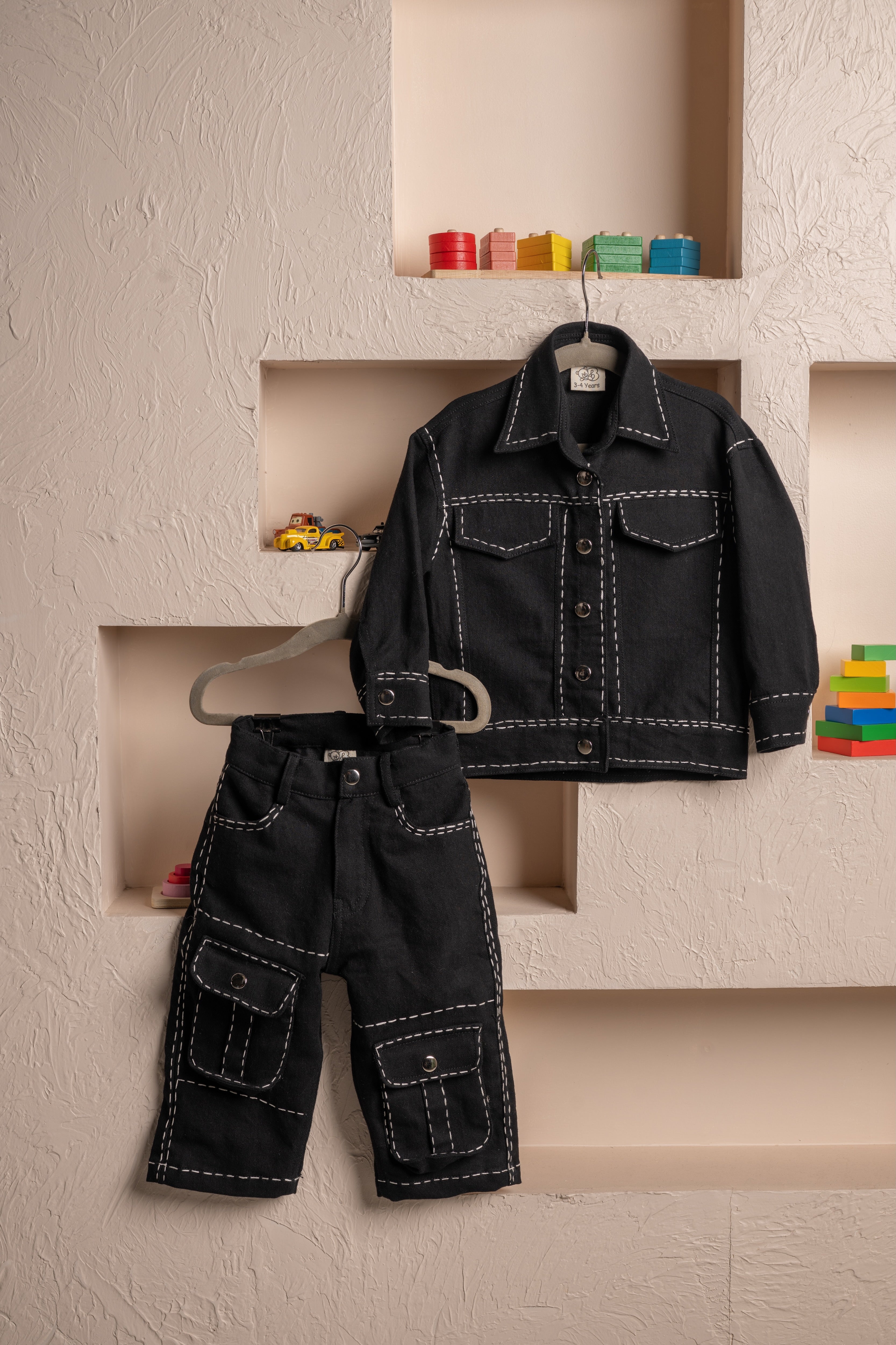 Luxury black toddler jacket and pants with white stitching displayed against textured beige wall with colorful wooden toys