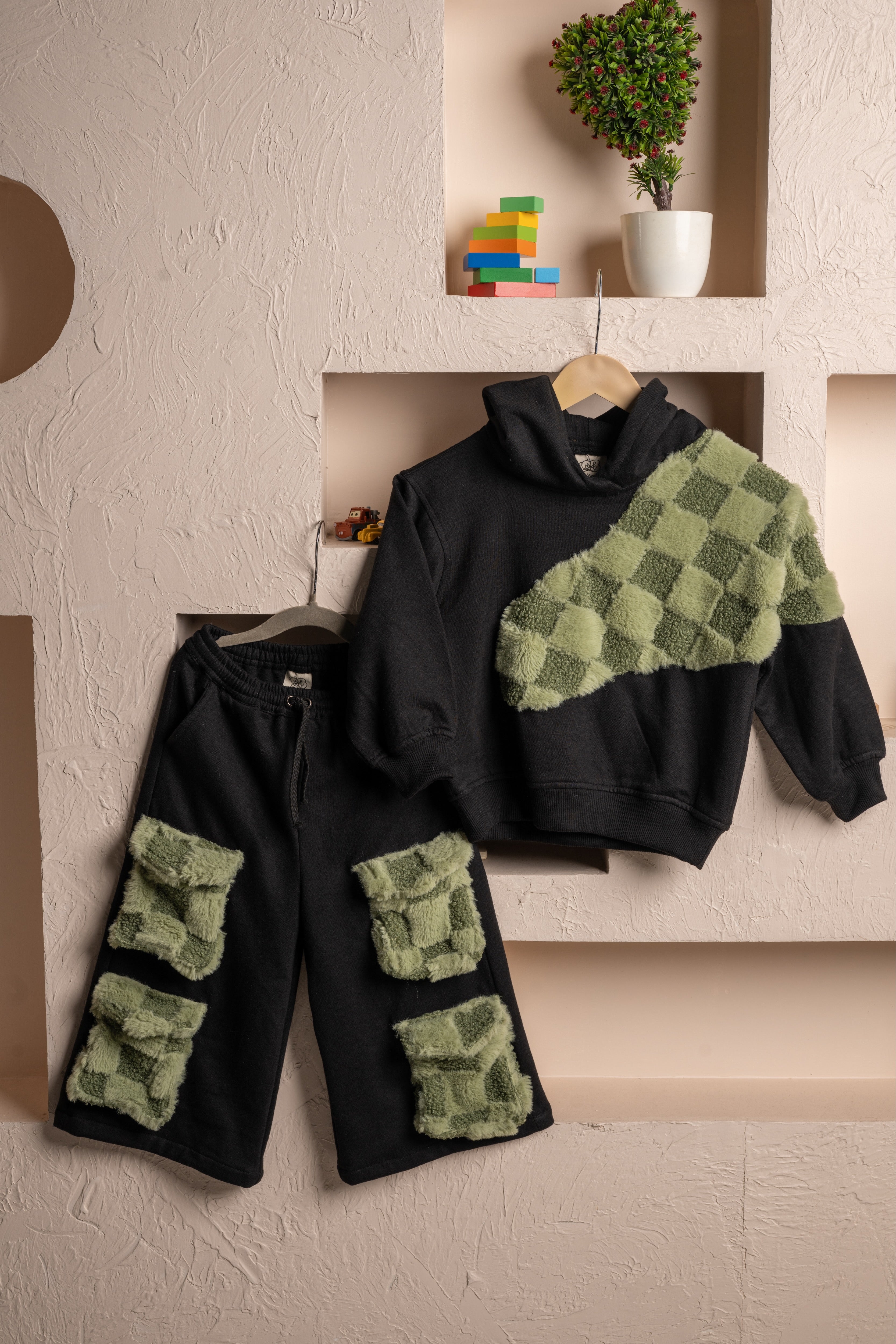 Black hoodie and pants set with textured green checkerboard patches displayed on beige textured wall