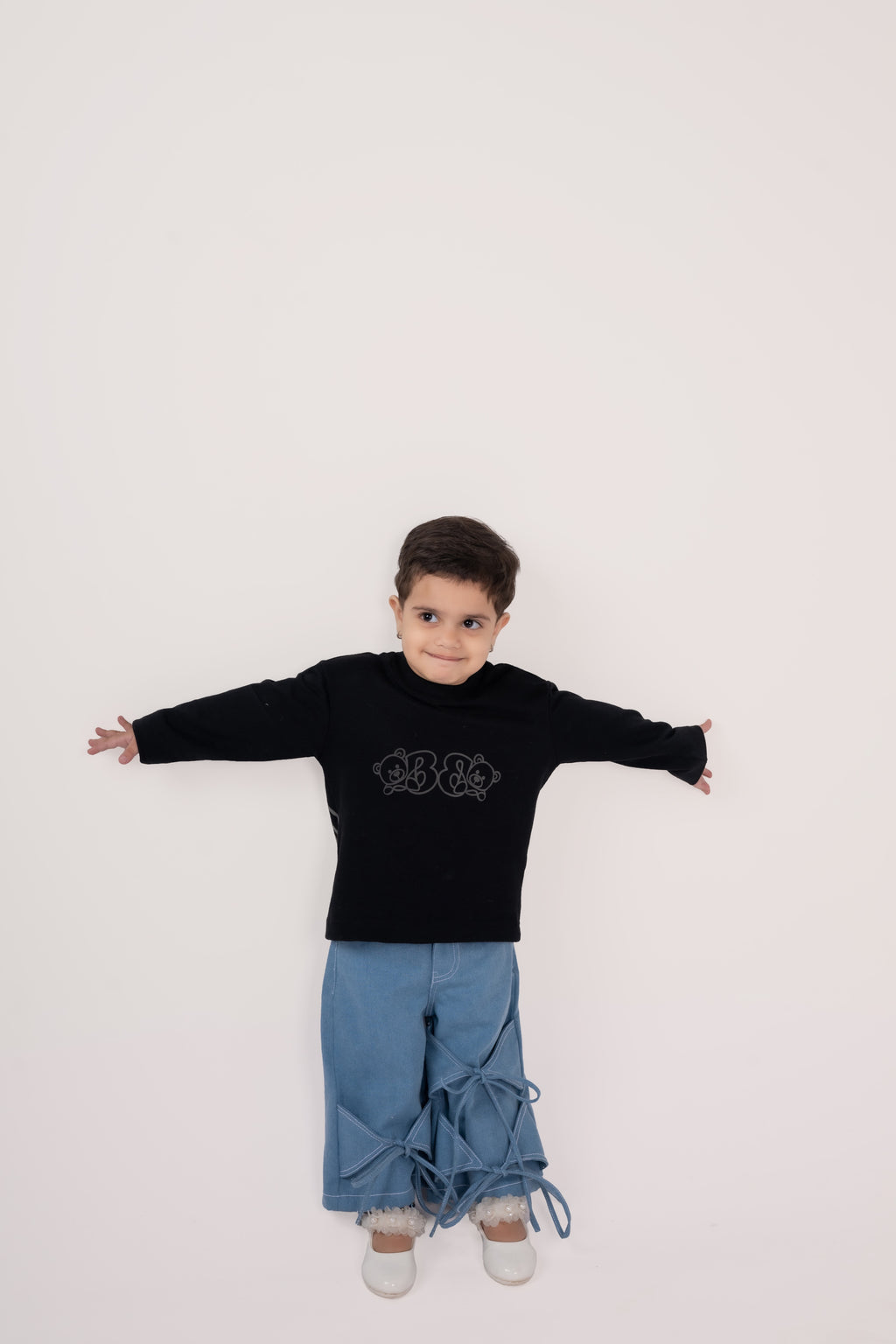Stylish toddler in black sweater and blue pants with bows, posing against a white background