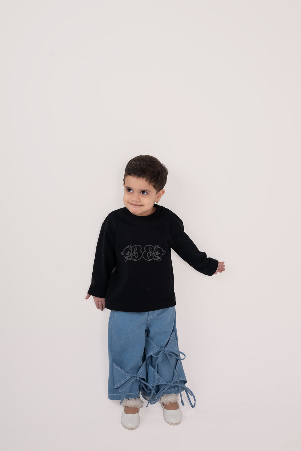 Stylish toddler in black sweater with bear design, blue wide-leg pants with bows, and white shoes