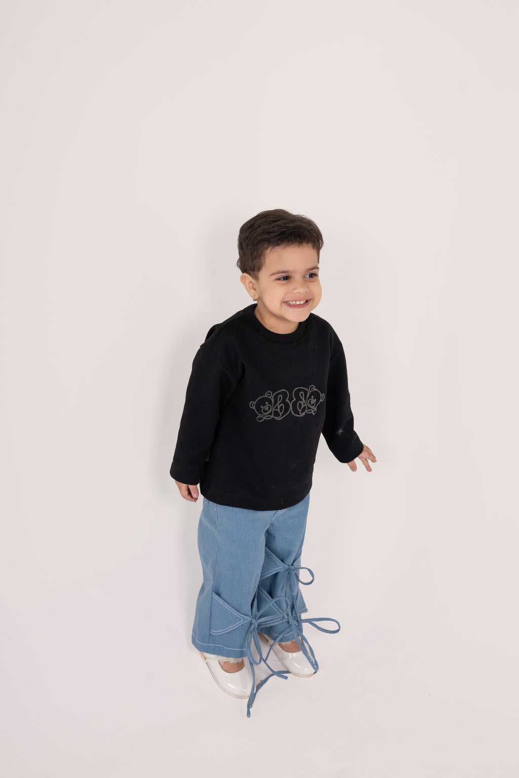 Smiling toddler in black bear-print sweater and stylish blue denim pants with tied bows, white shoes
