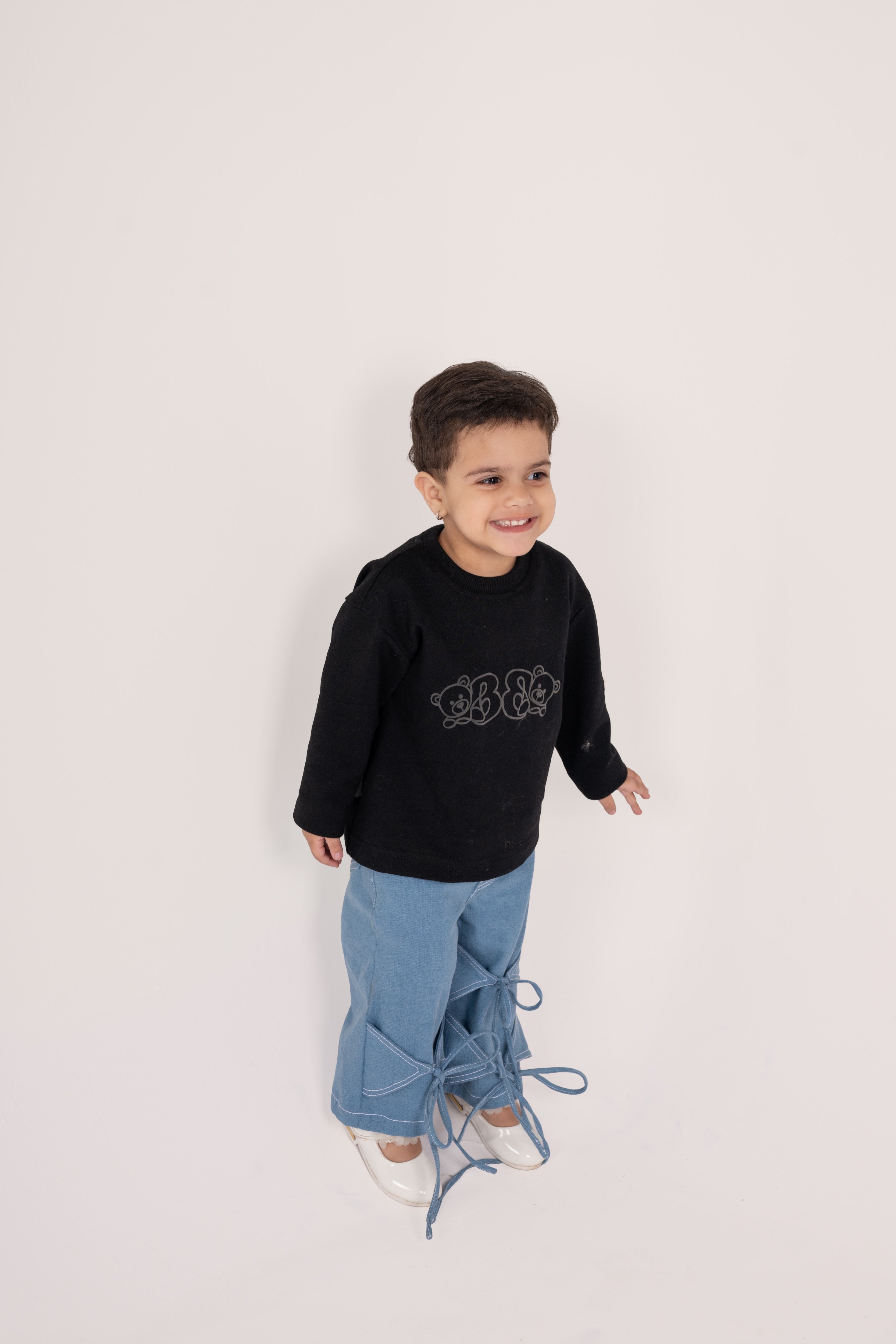 Smiling toddler in black sweatshirt with bear design and stylish blue denim pants with bow details
