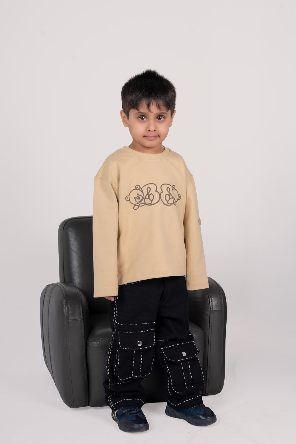 Young boy standing in stylish beige sweater with bear print and black cargo pants in modern setting