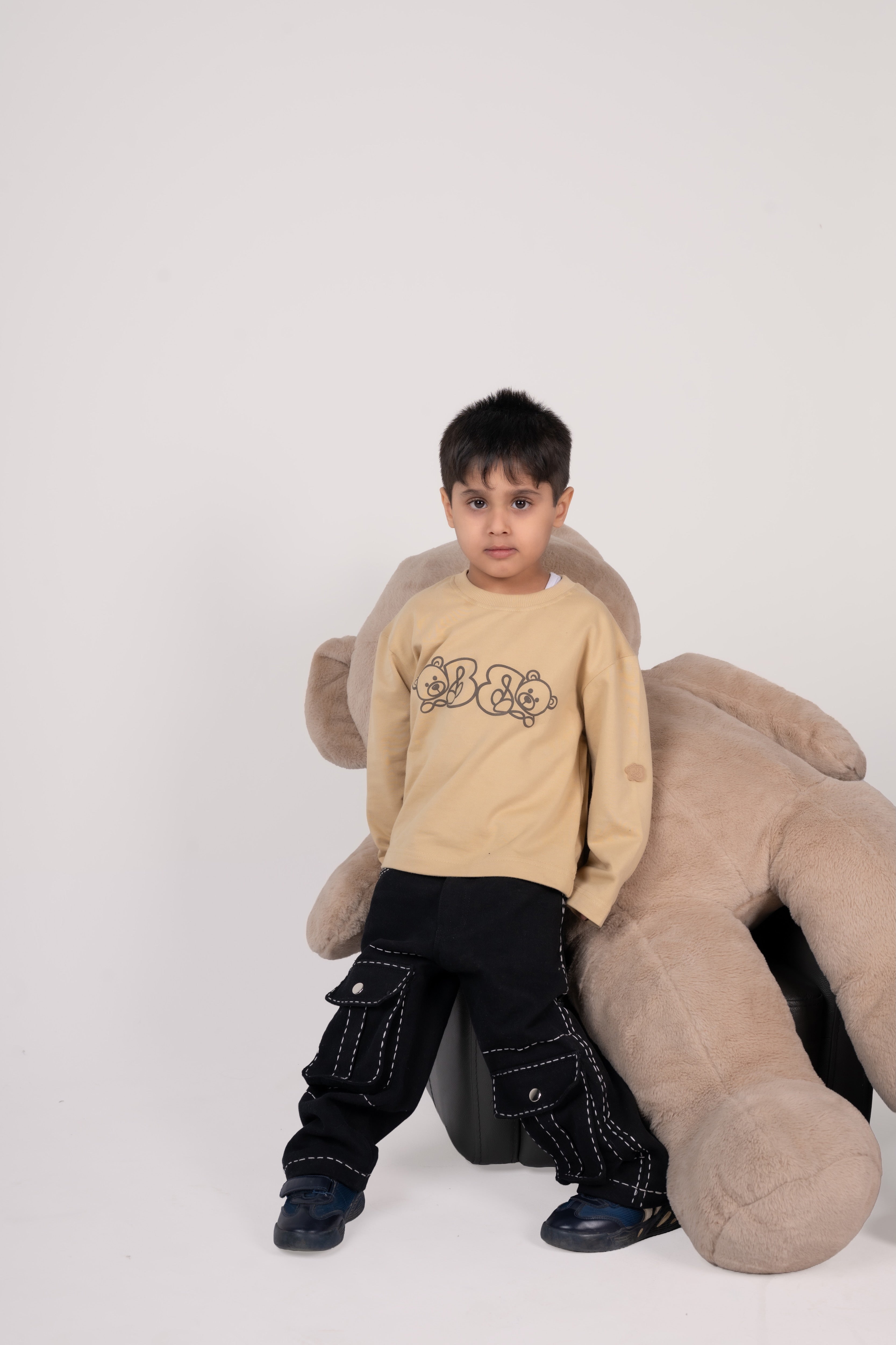 Stylish young boy in beige teddy bear sweatshirt and black cargo pants posing with oversized plush bear
