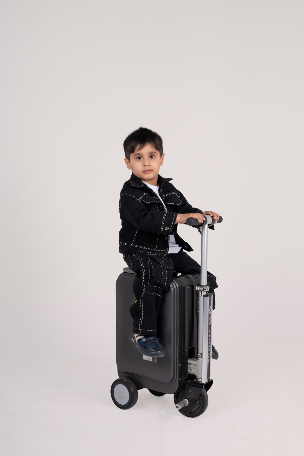 Stylish young boy in black outfit sitting on modern scooter luggage against plain backdrop