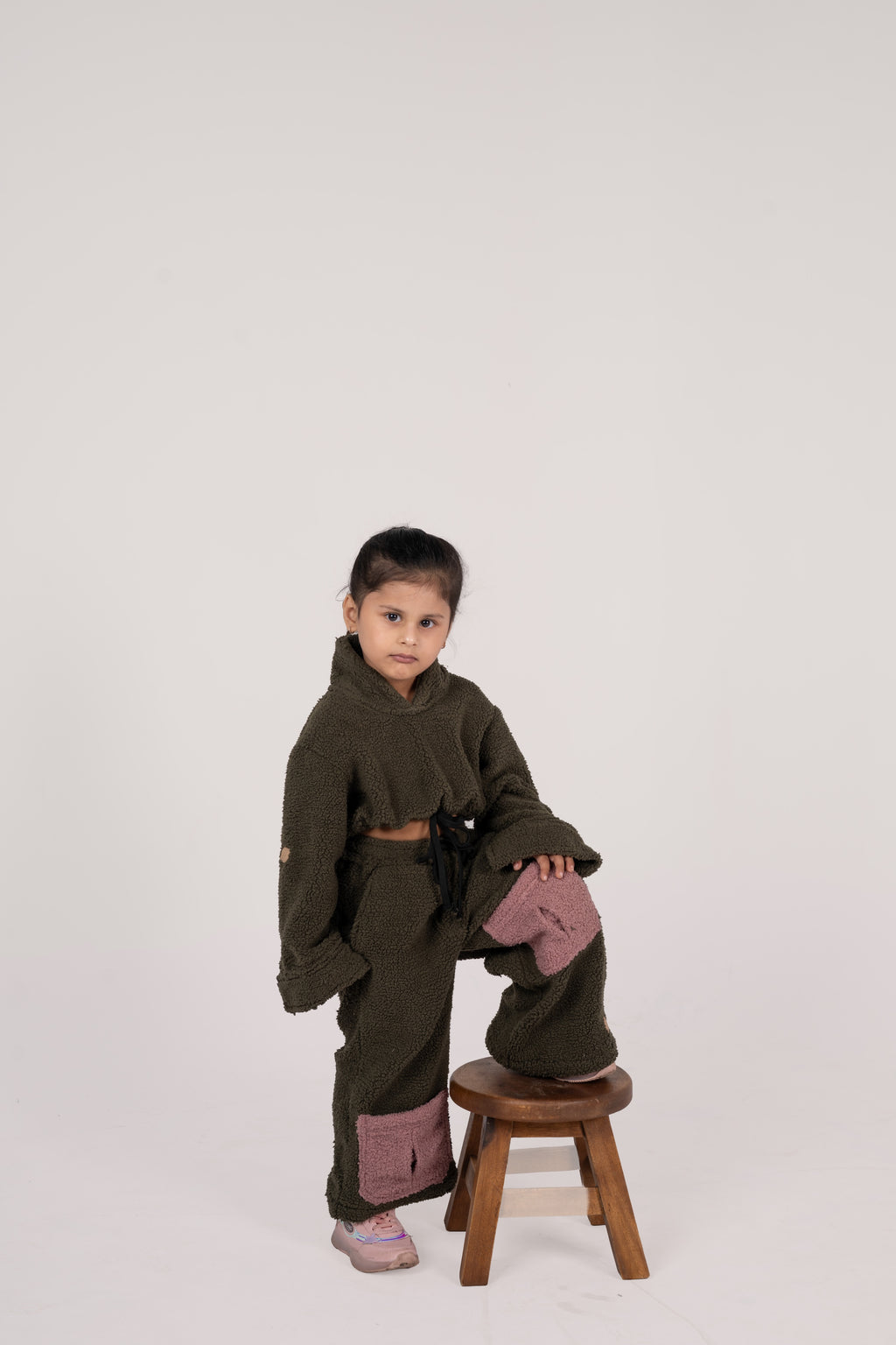 Stylish young girl in luxurious olive green fleece set with pink patches posing on wooden stool