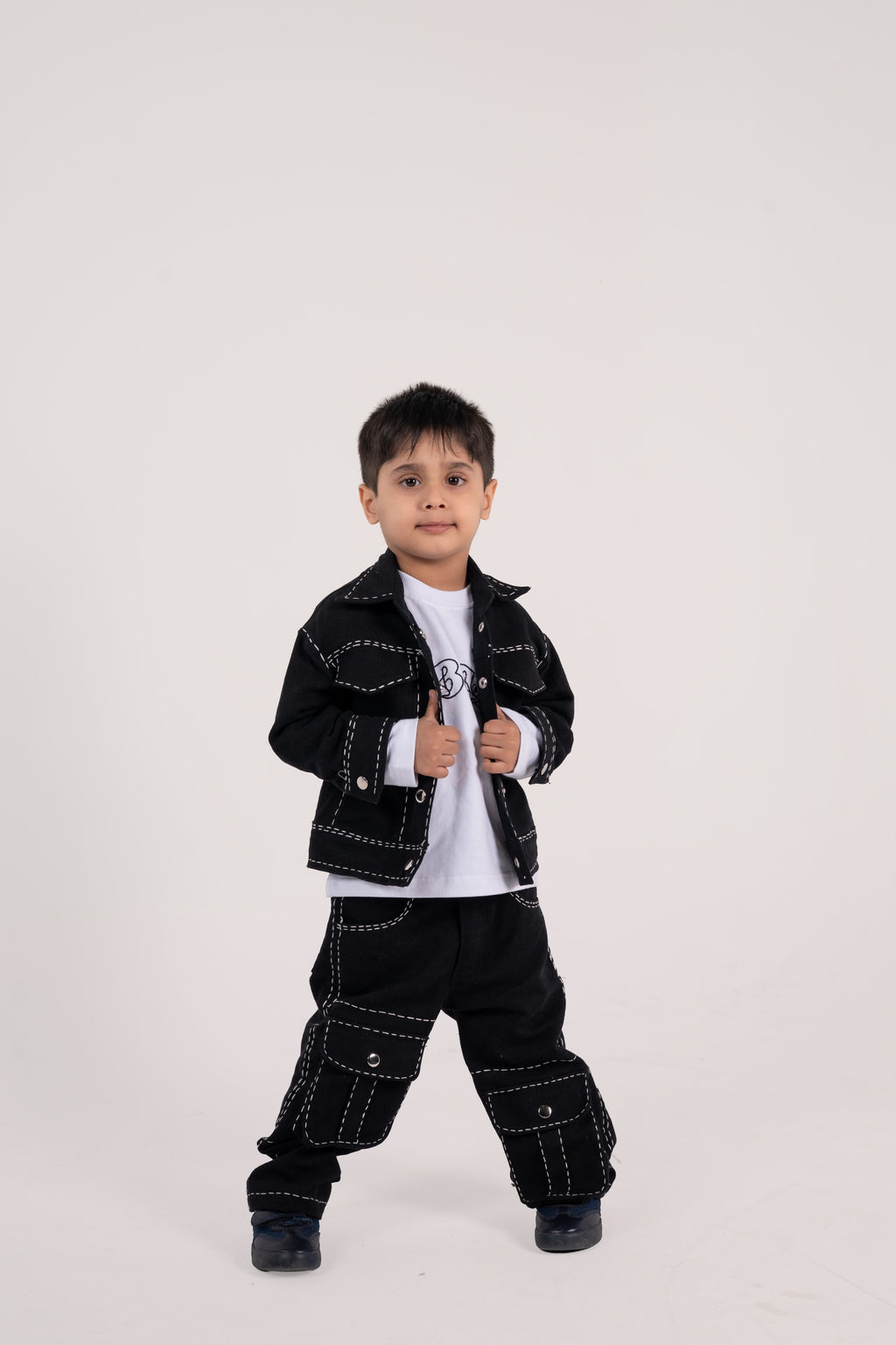 Young boy in stylish black jacket and cargo pants with white stitching on white background