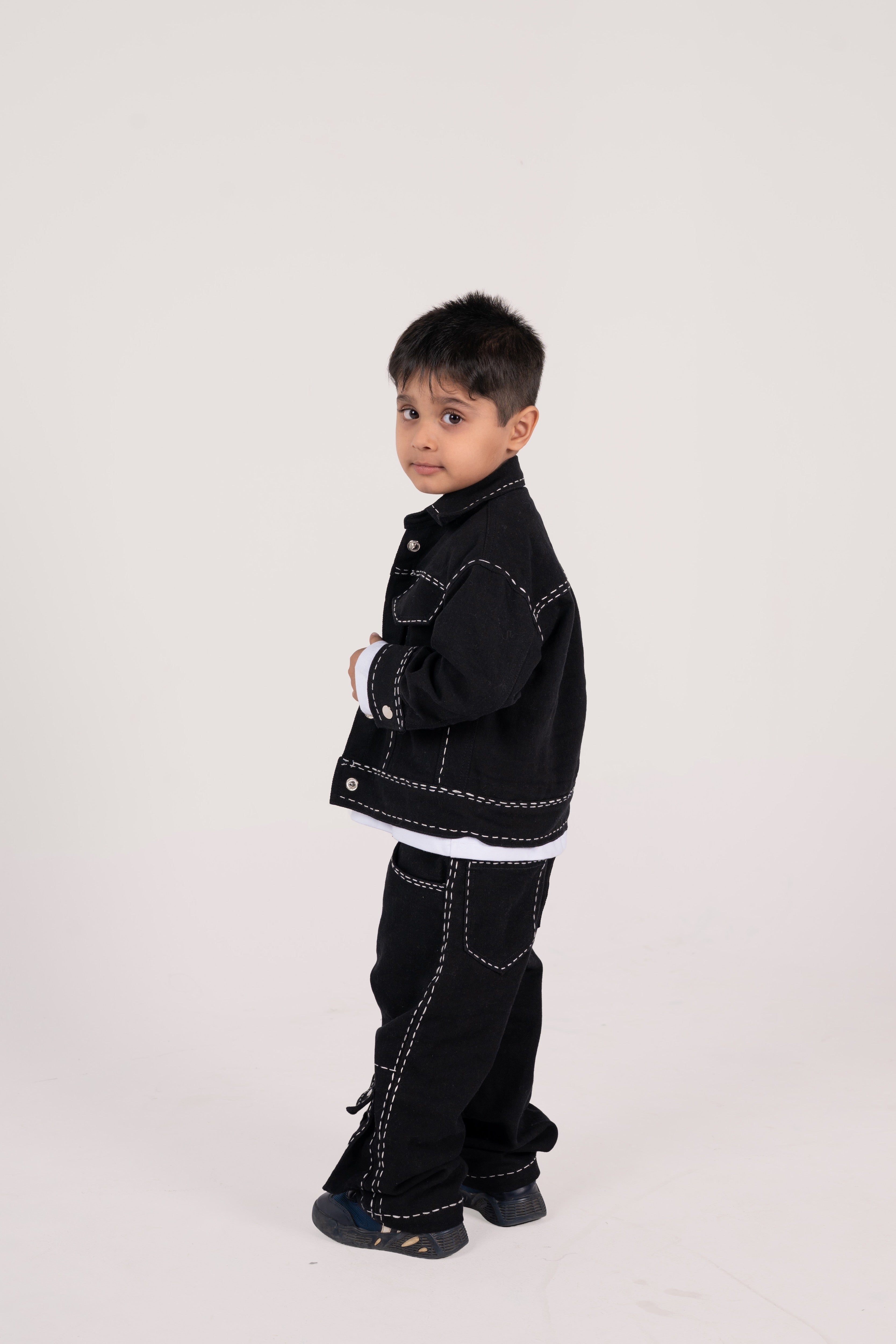 Young boy in stylish black denim jacket and pants with white stitching on plain background