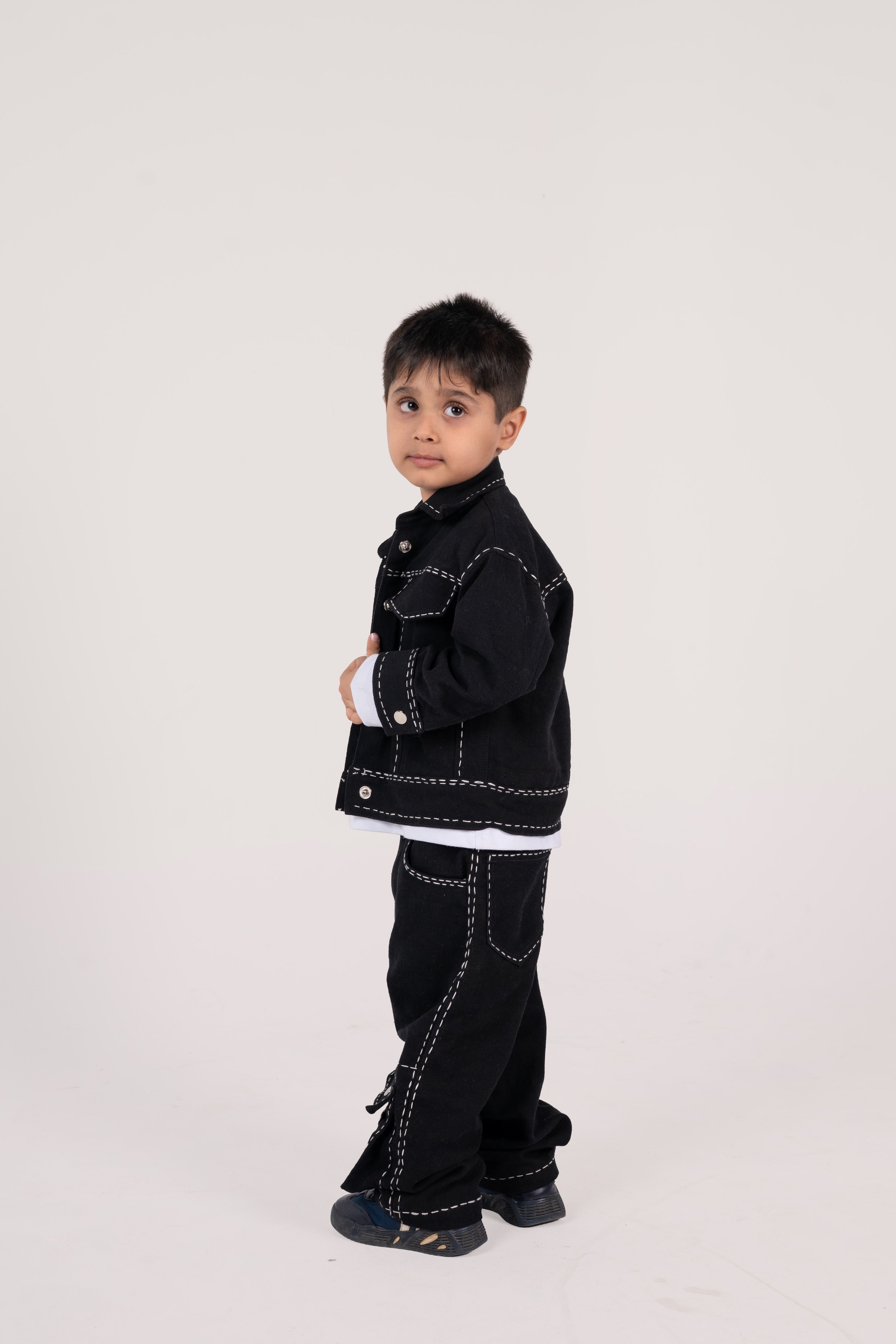 Young boy in stylish black denim outfit with white stitching, posing against a light background