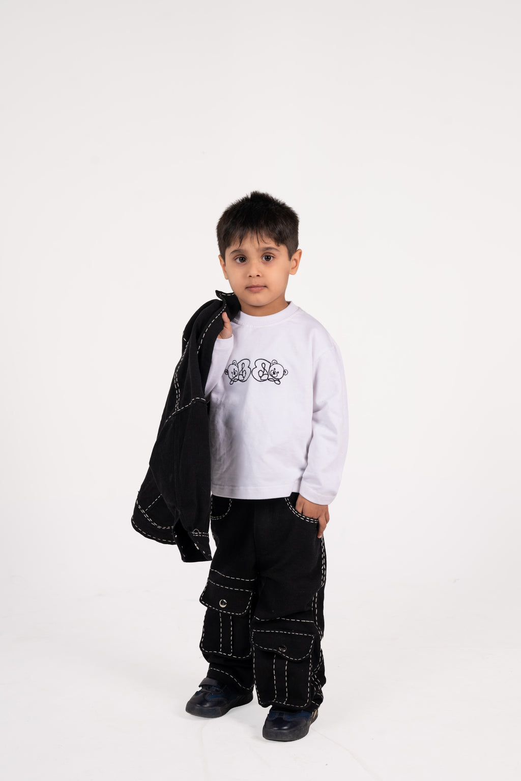 Young boy in stylish black cargo pants and white long sleeve shirt holding black jacket in studio
