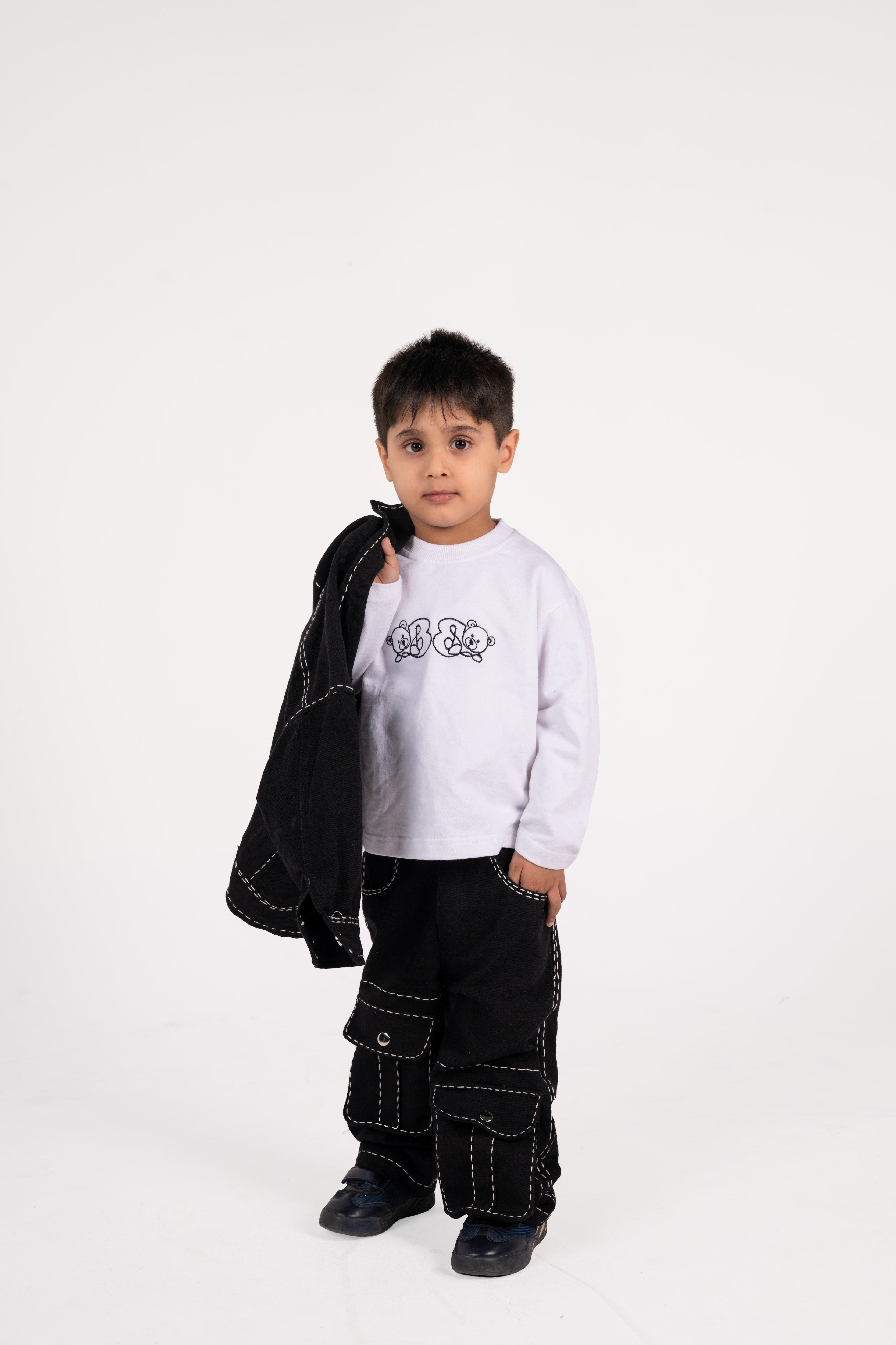 Young boy in stylish black cargo pants and white long sleeve shirt holding black jacket in studio