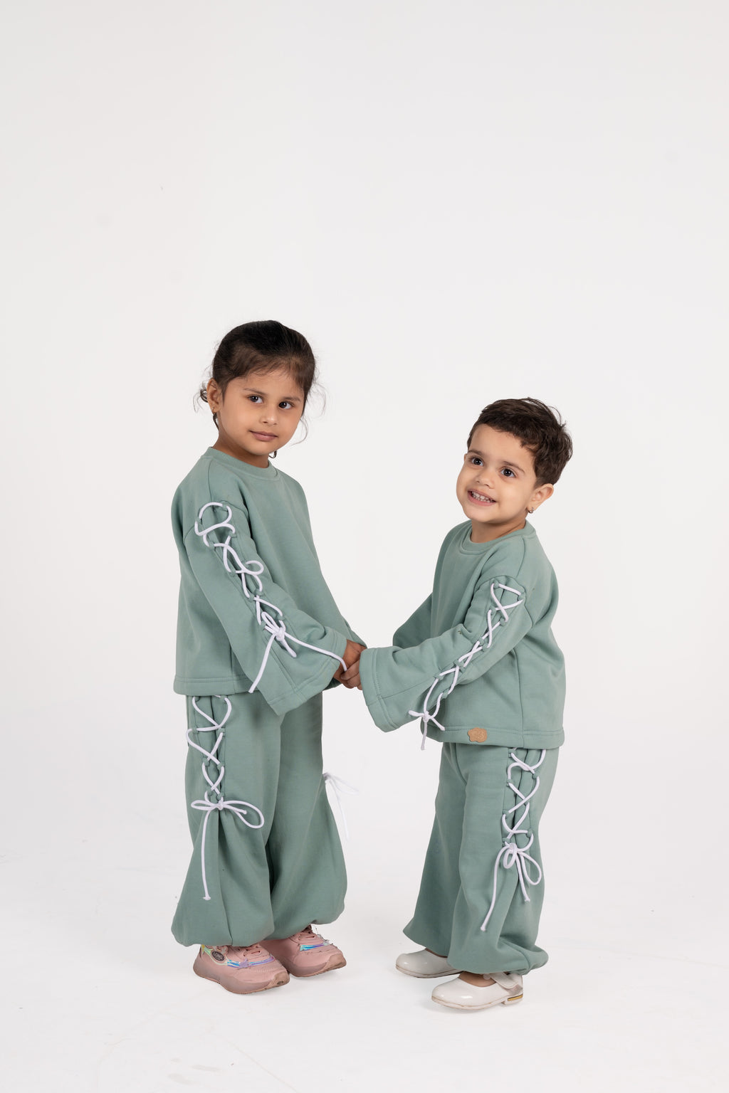 Two children wearing matching sage green laced lounge sets holding hands against white background