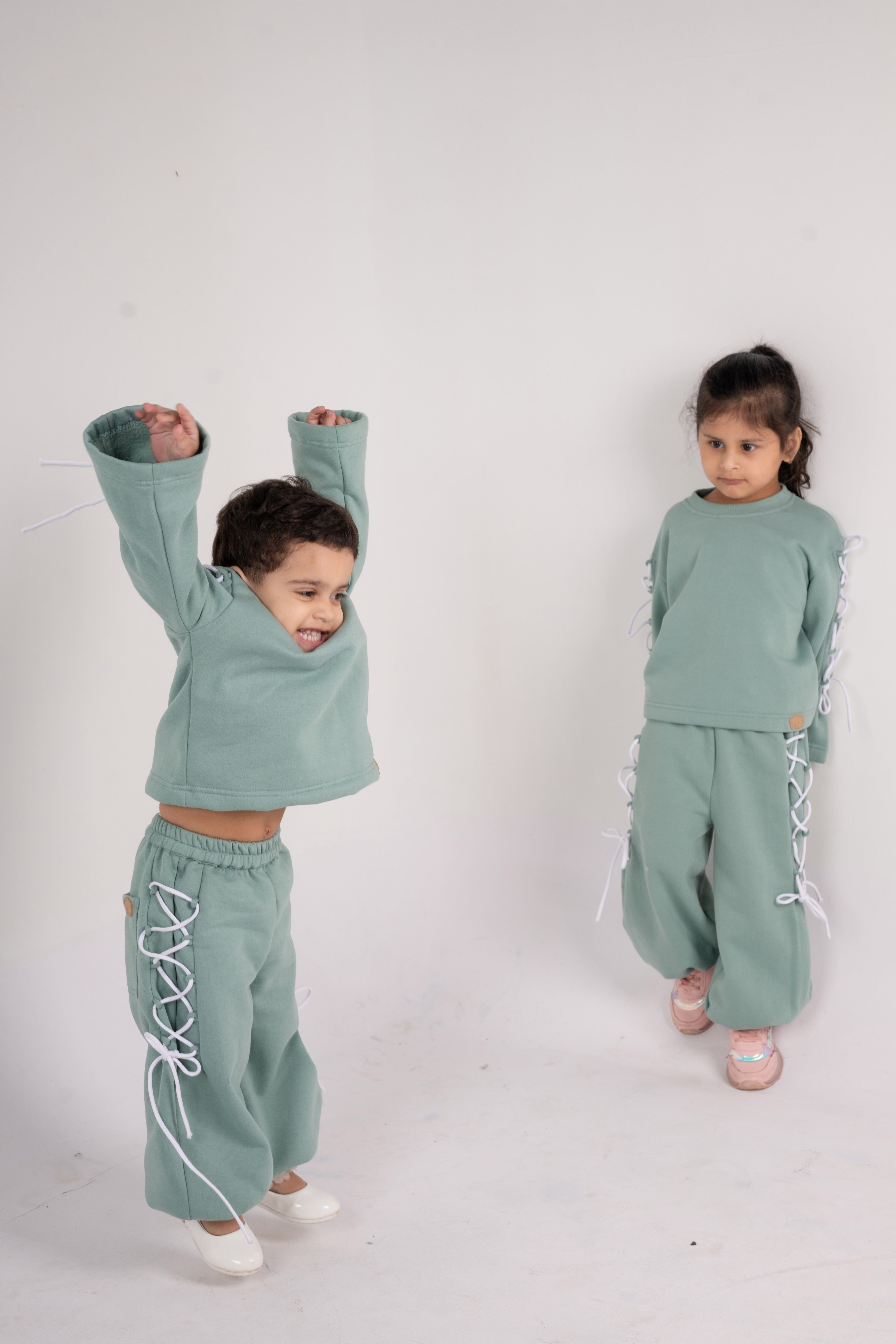 Two children in pastel green matching loungewear sets with white lace-up details on sleeves and pants against plain background