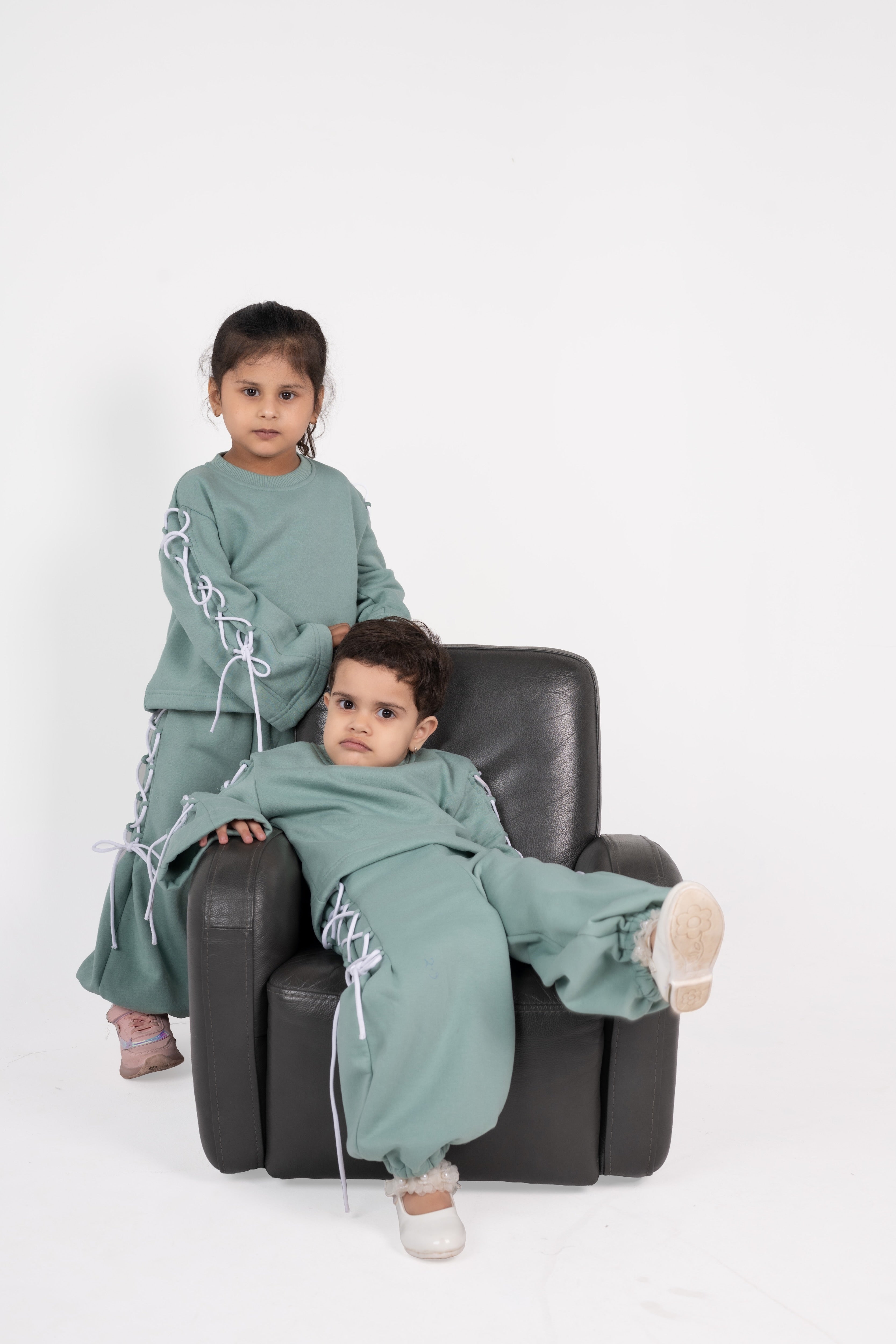 Two children in stylish sage green loungewear with white lace-up details, sitting on a black leather armchair