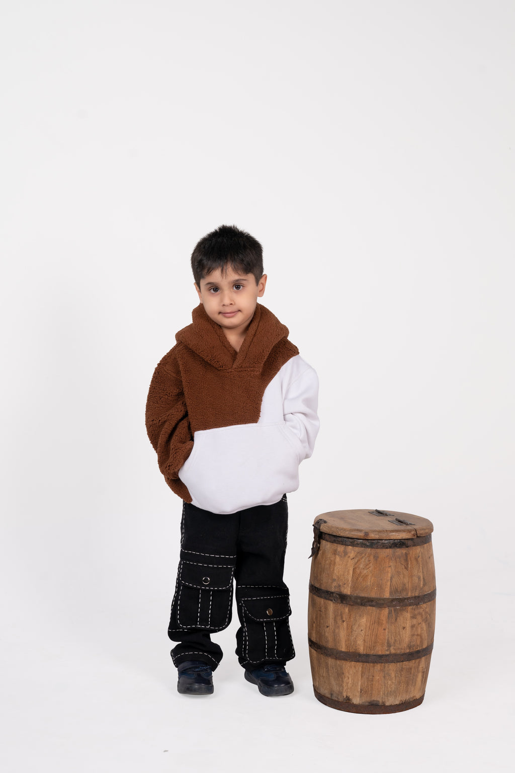 Young boy in stylish brown and white hoodie with black cargo pants standing beside a rustic wooden barrel