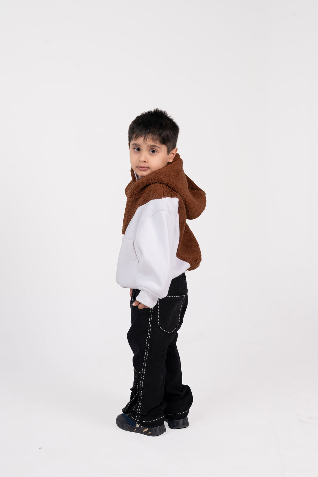 Stylish young boy in white sweatshirt with brown hood and black pants against white background