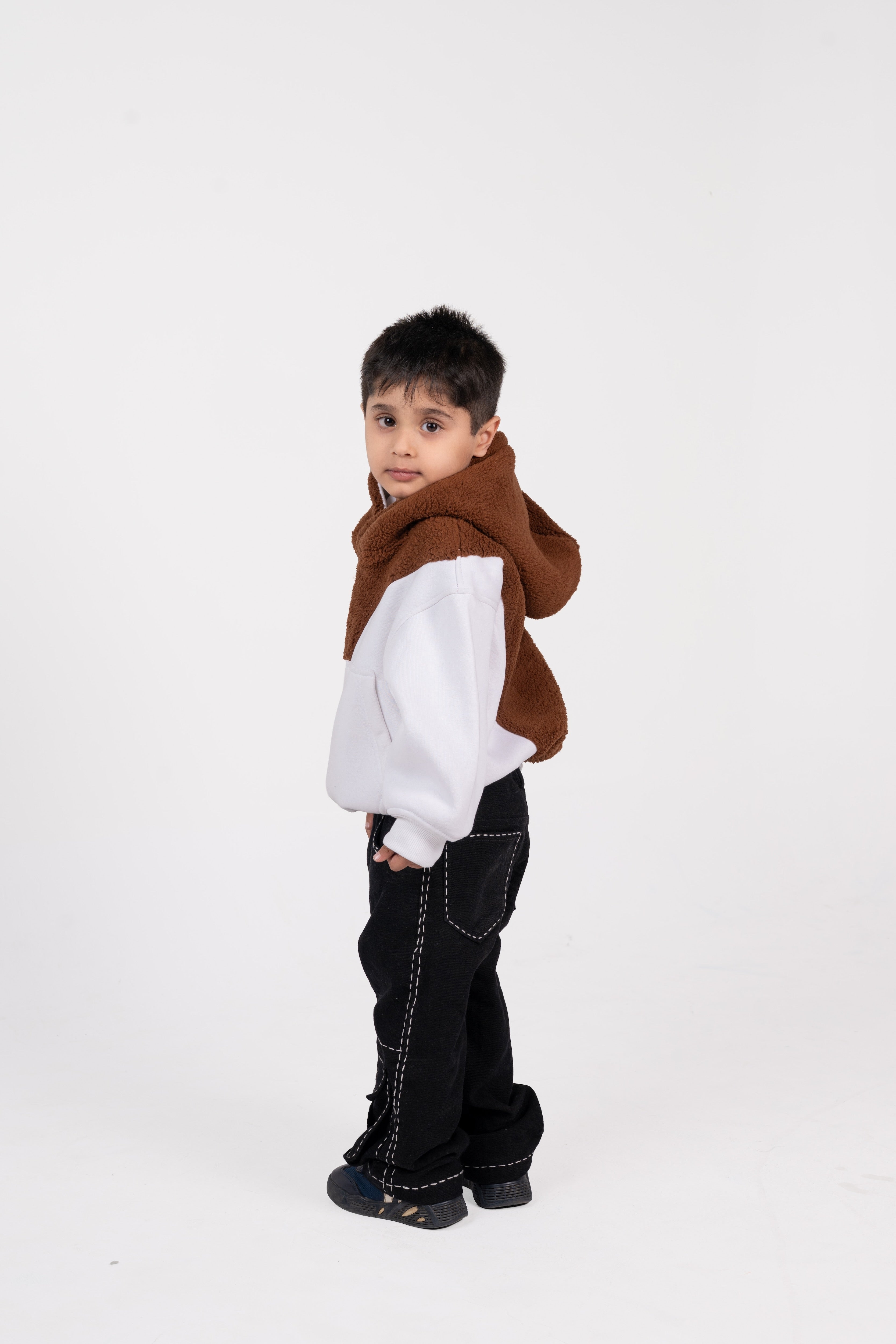 Stylish young boy in white sweatshirt with brown hood and black pants against white background