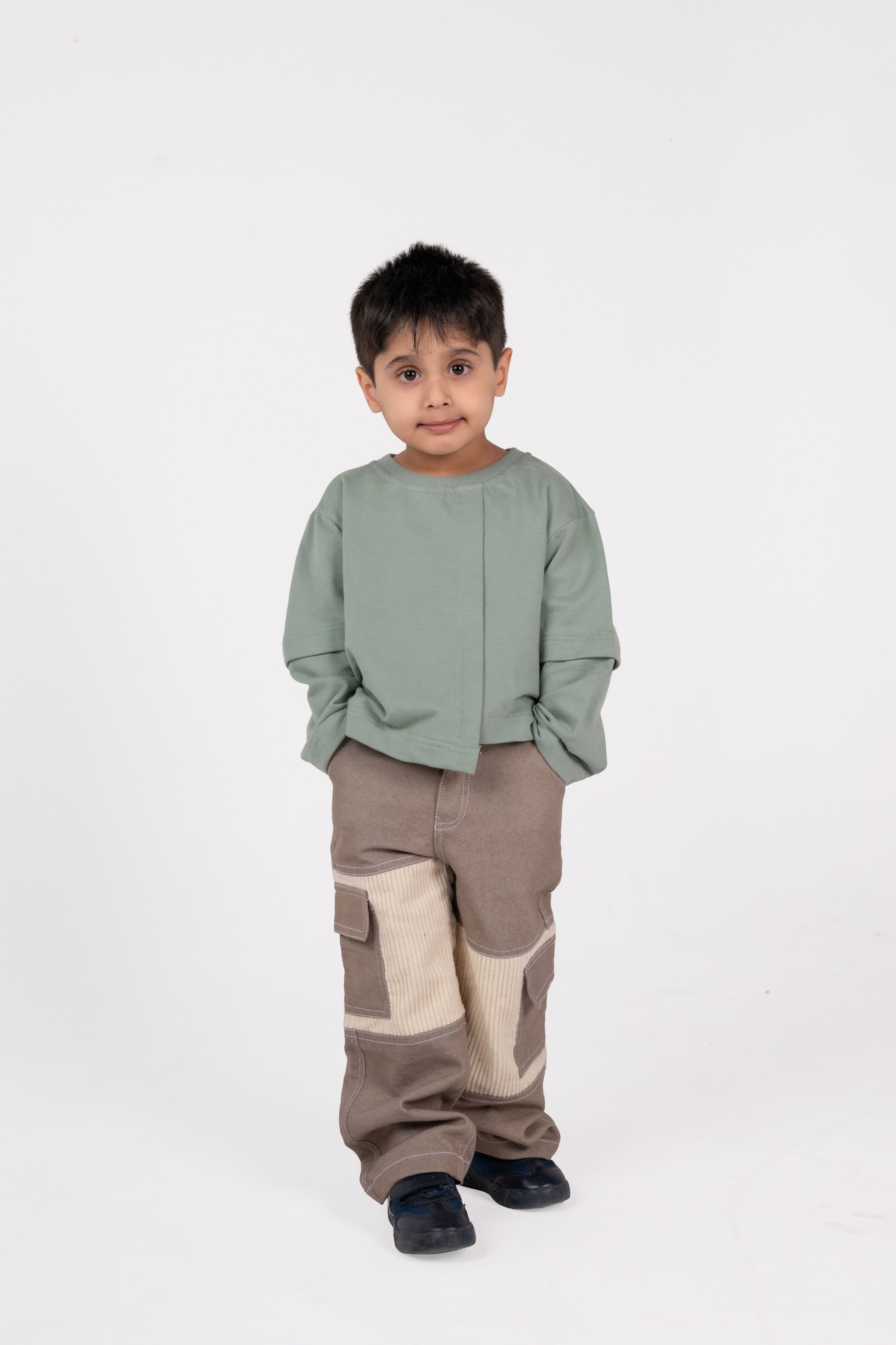 Stylish young boy in sage green sweatshirt and patchwork beige cargo pants against white background