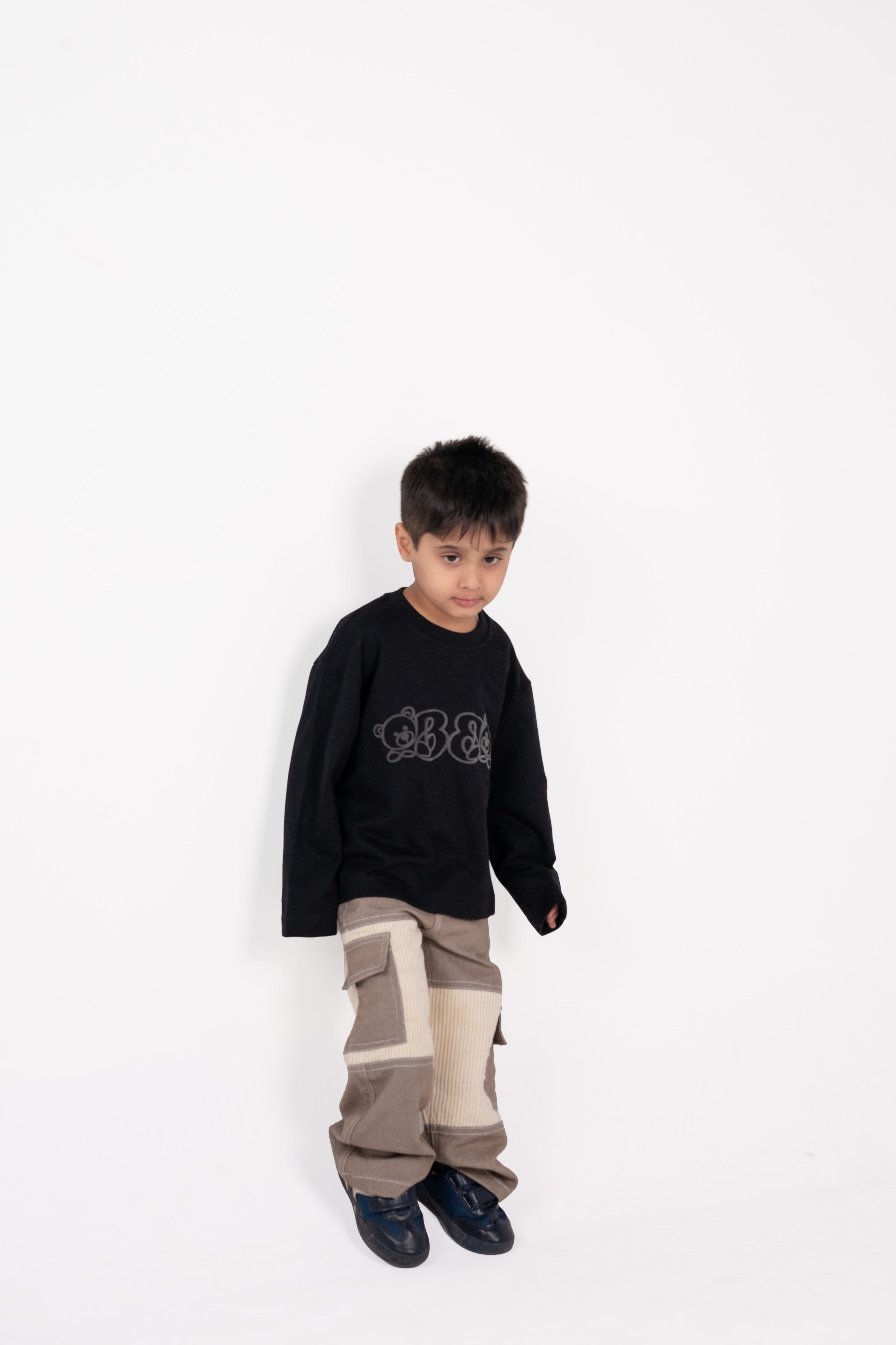 Young boy in stylish black graphic sweater and beige patchwork pants against white background