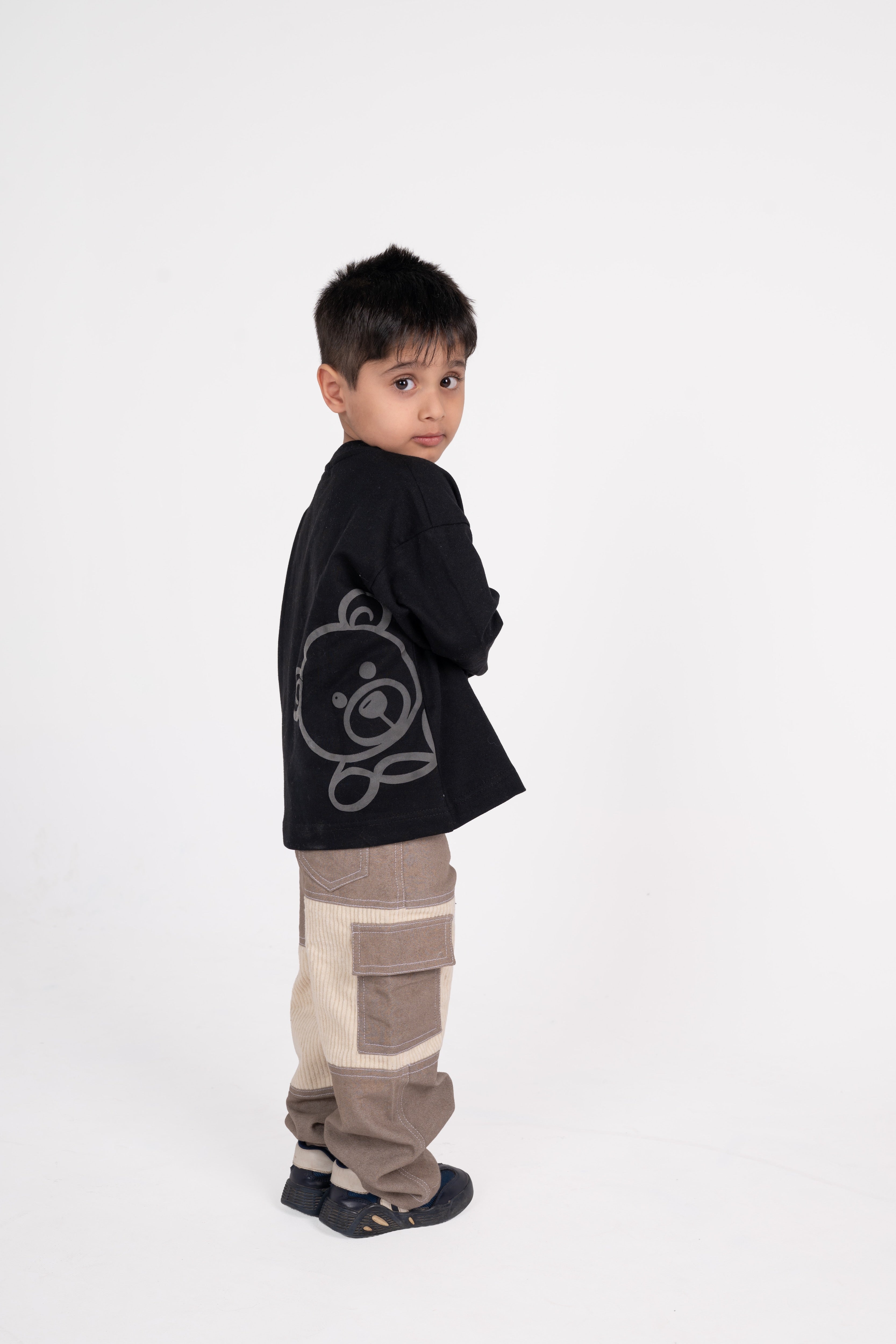 Stylish young boy in black graphic sweater and beige patchwork pants posing against white backdrop