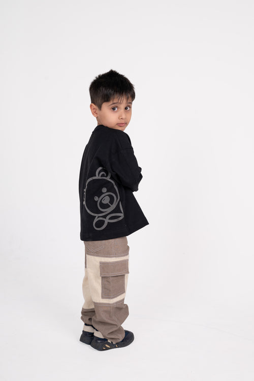 Stylish young boy in black graphic sweater and beige patchwork pants posing against white backdrop