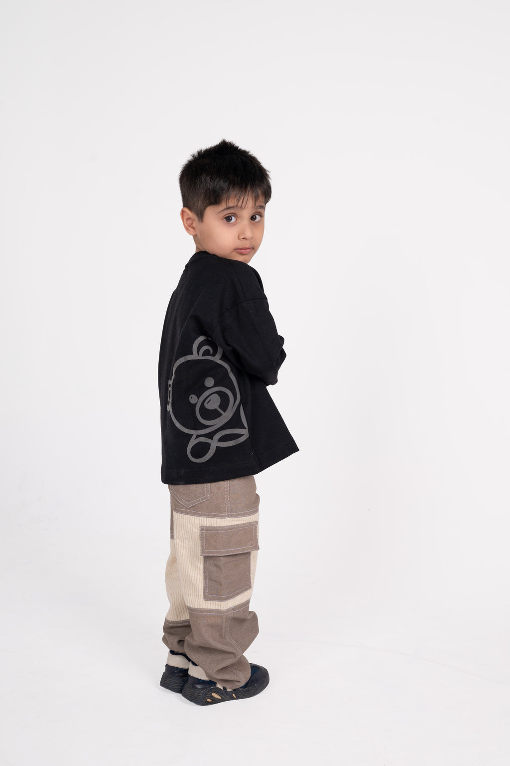 Young boy in stylish black sweater with bear print and patchwork cargo pants on white backdrop