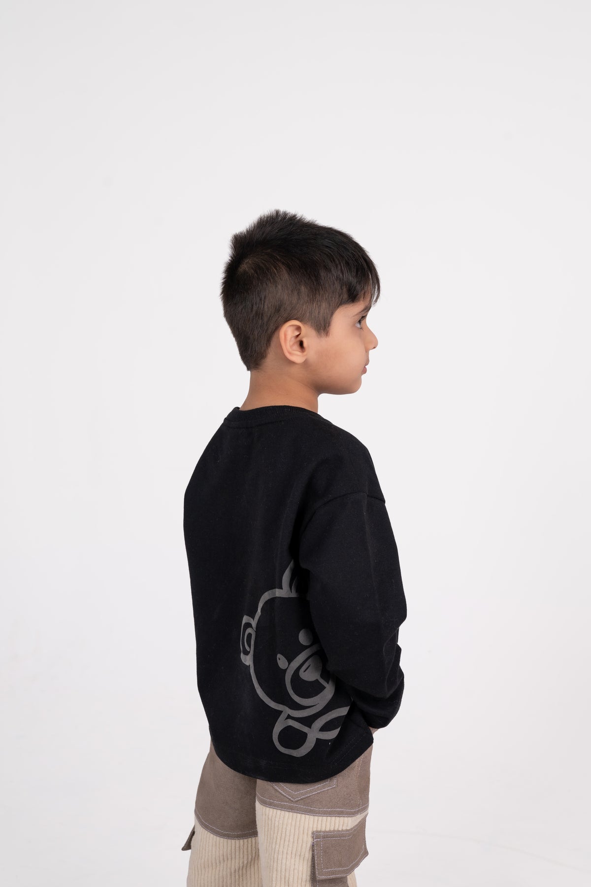 Stylish young boy in black sweater with teddy graphic and beige cargo pants on white background