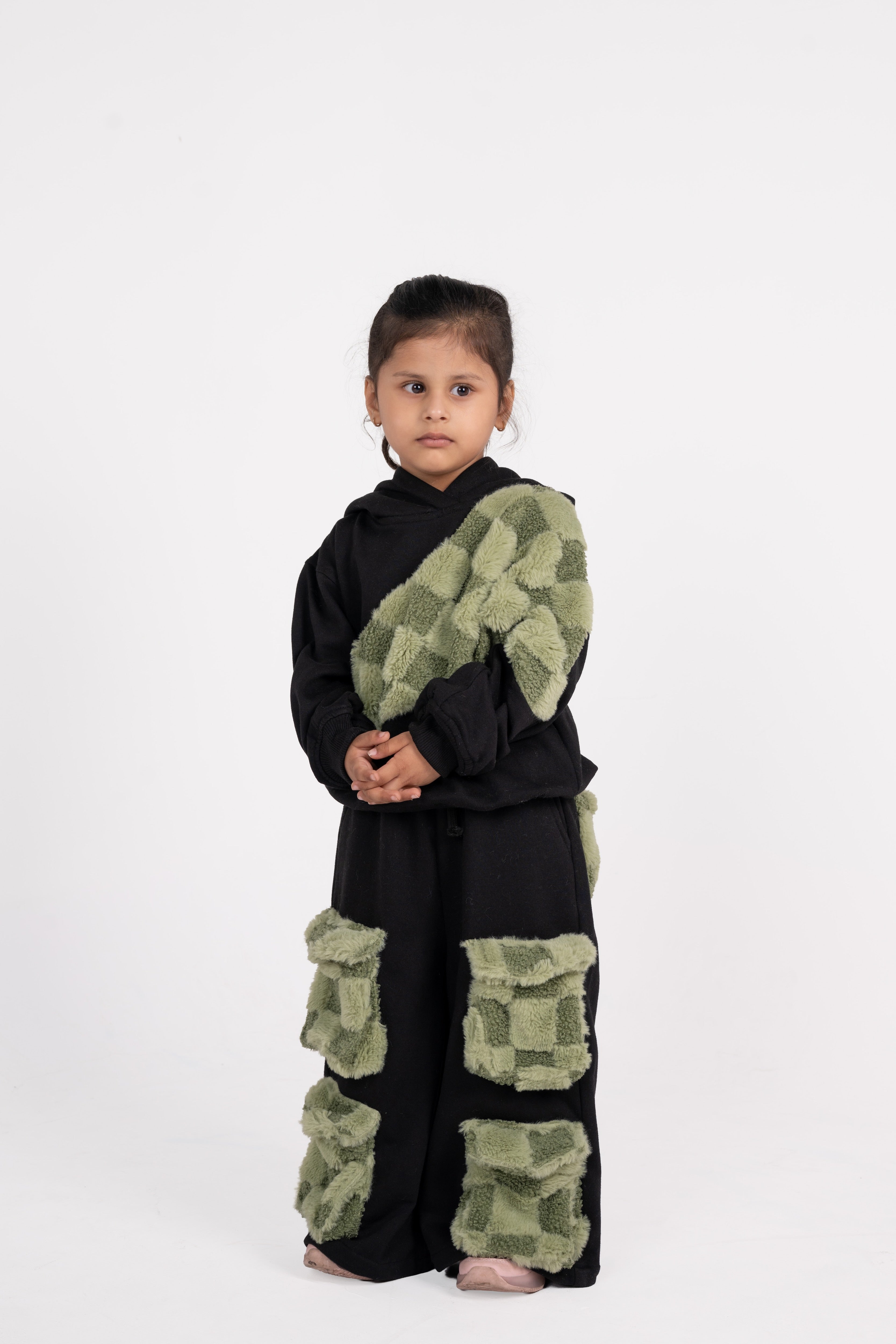 Young girl in black outfit featuring green checkered faux fur accents on pants and shoulder sash