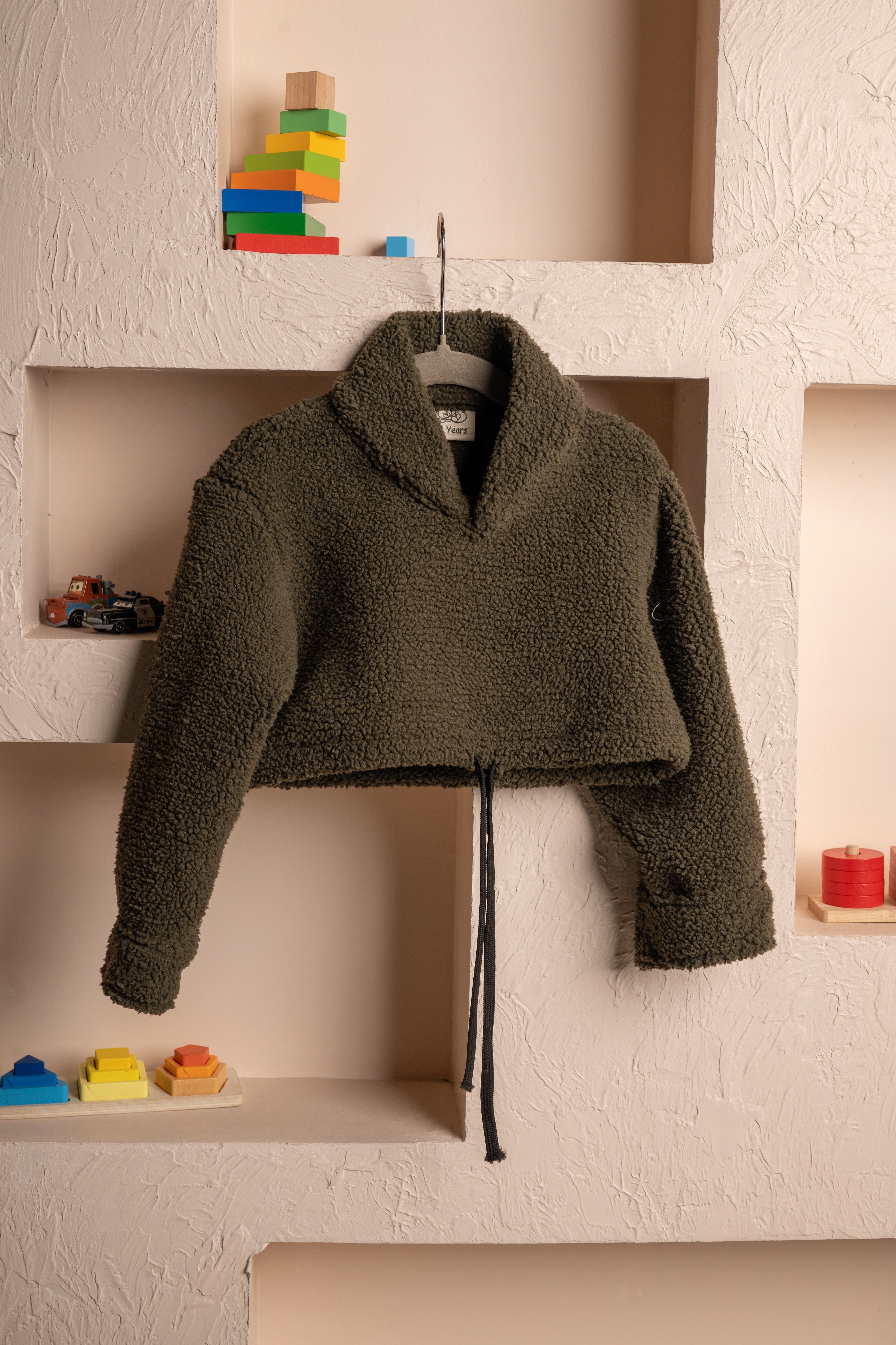 Luxurious olive green cropped sherpa fleece jacket with shawl collar displayed on textured beige wall niches