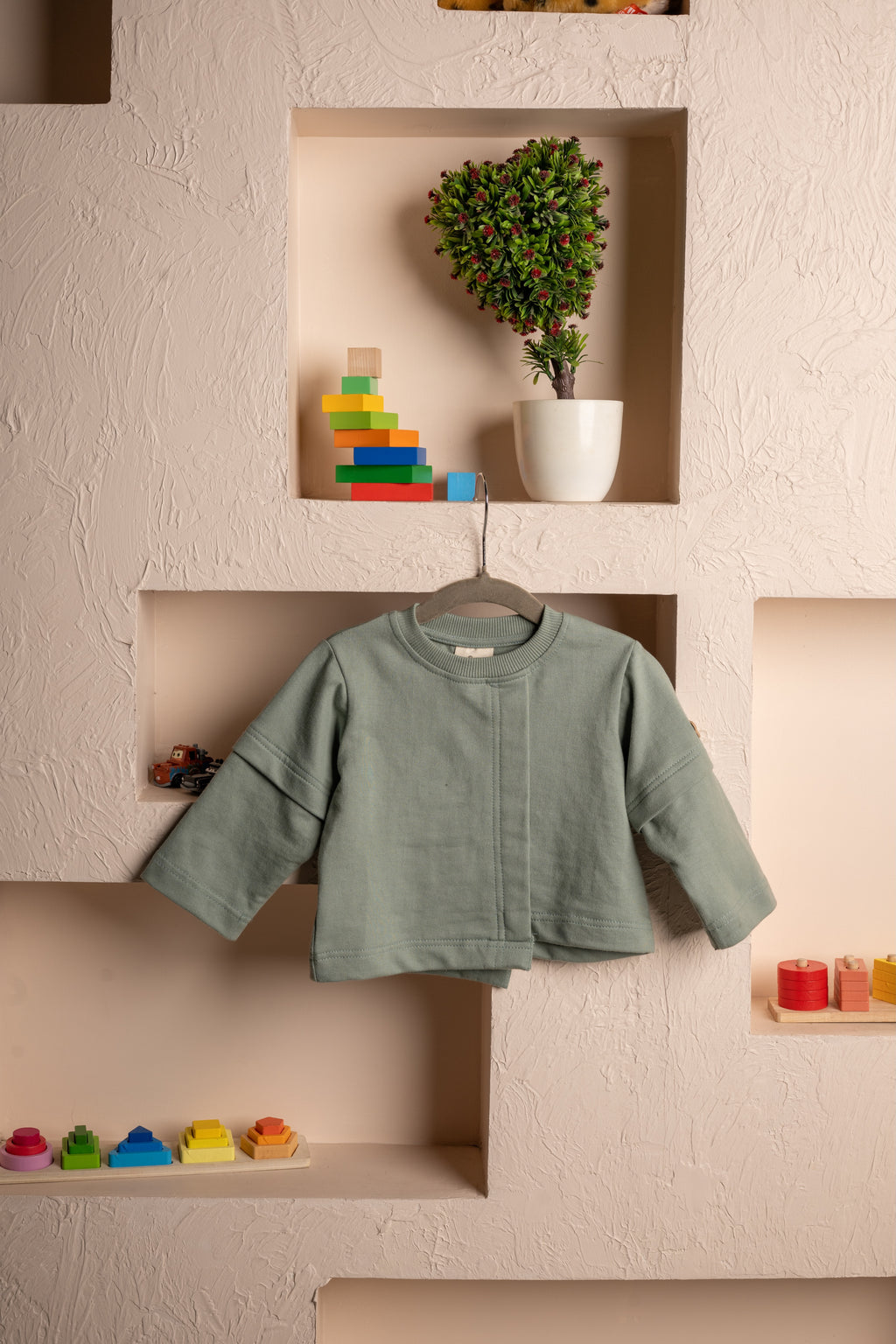 Luxury kids sage green jacket hanging on textured wall with colorful wooden toys and potted topiary