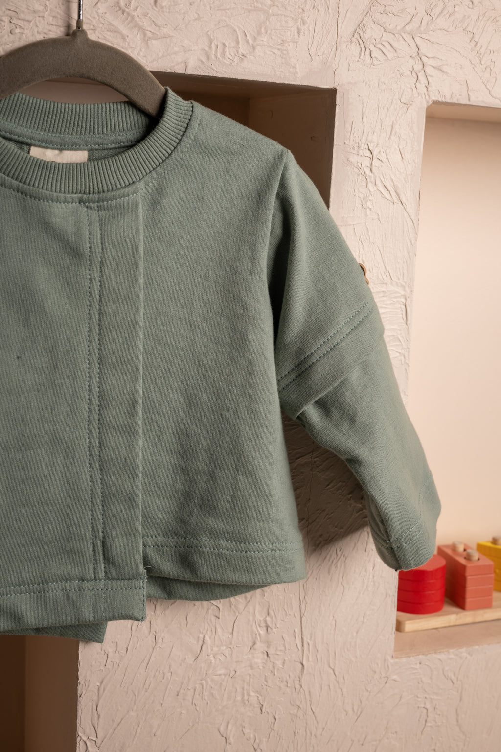 Soft sage green baby cardigan hanging against a textured beige wall with wooden blocks in niche