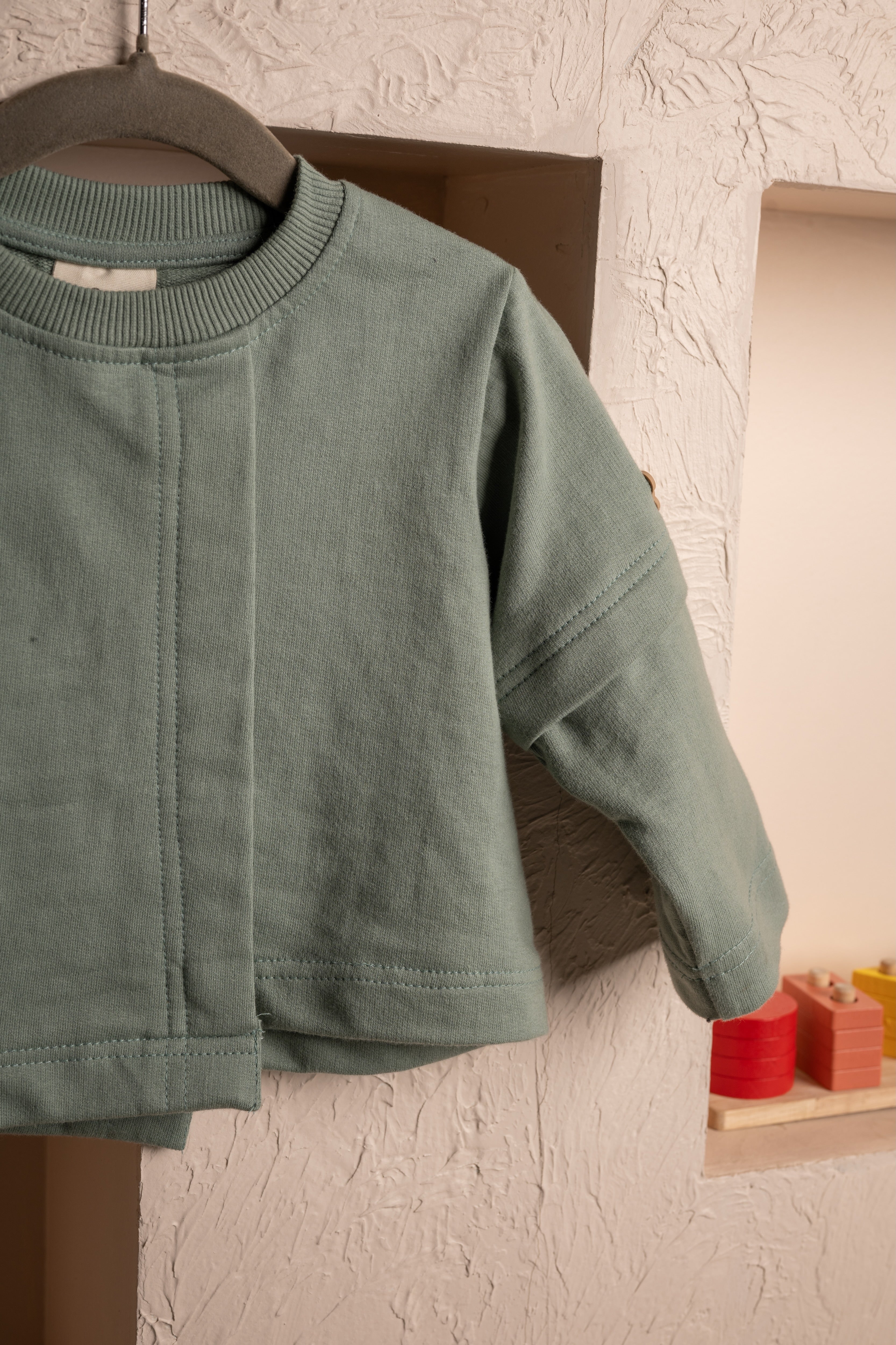 Soft sage green baby cardigan hanging against a textured beige wall with wooden blocks in niche