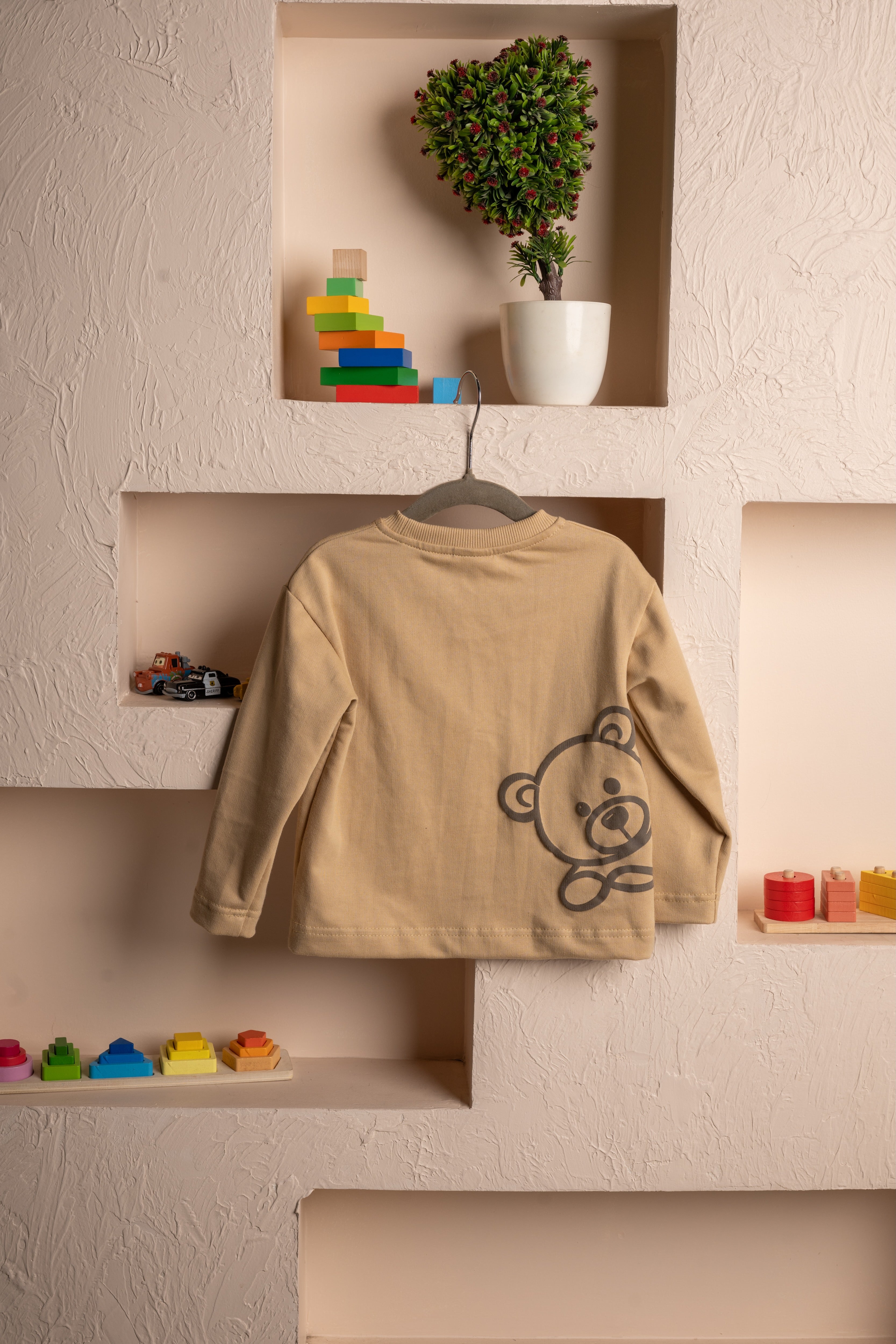 Elegant beige baby sweater with a teddy bear print hanging on textured wall shelves with colorful children's toys