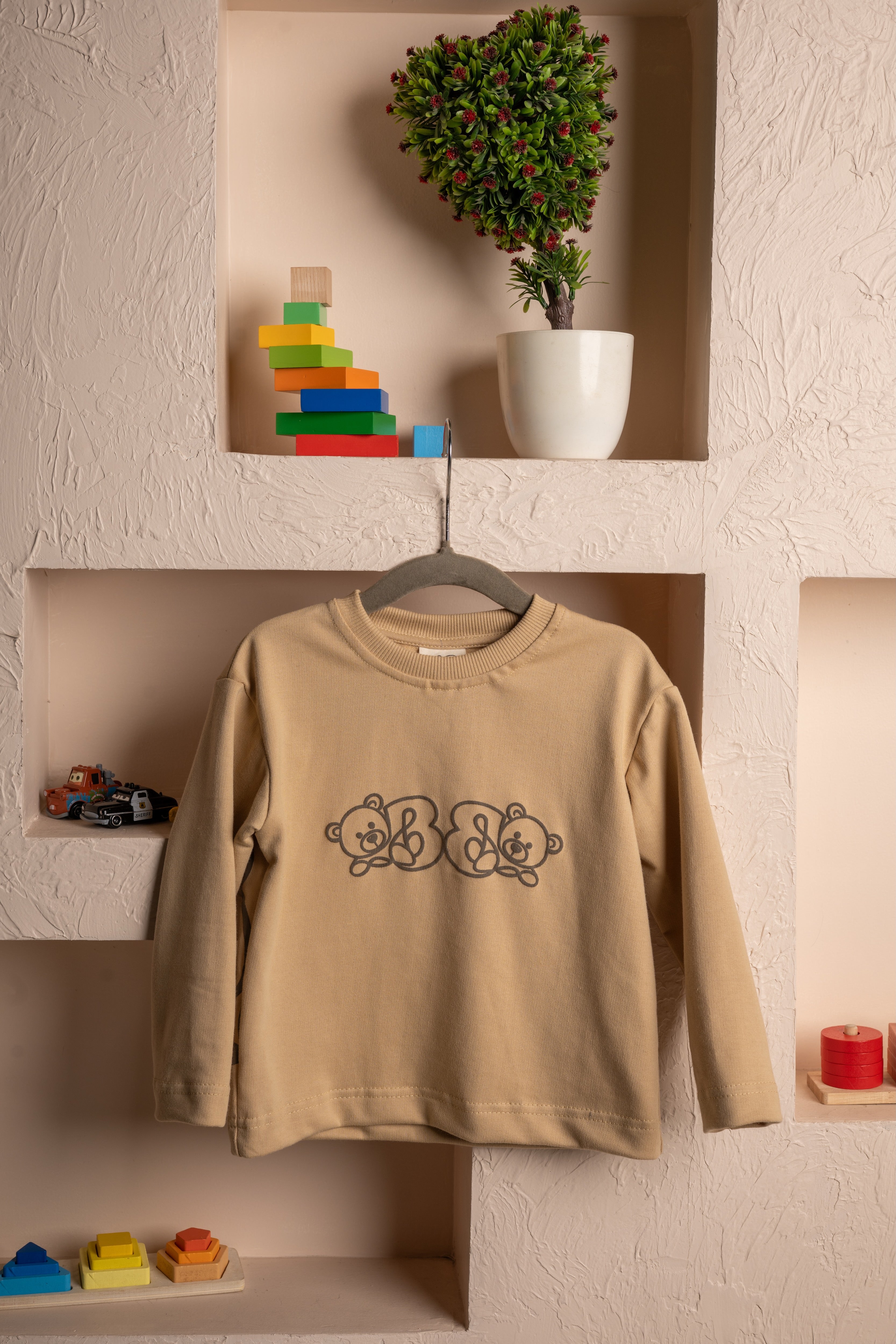 Elegant beige children's sweatshirt with teddy bear design hanging on textured wall with colorful toys and potted plant