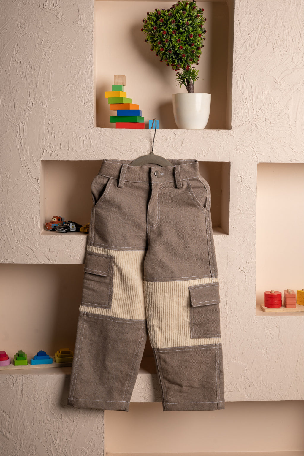 Elegant brown and beige patchwork kids cargo pants hanging on a textured wall with decorative shelves and toys