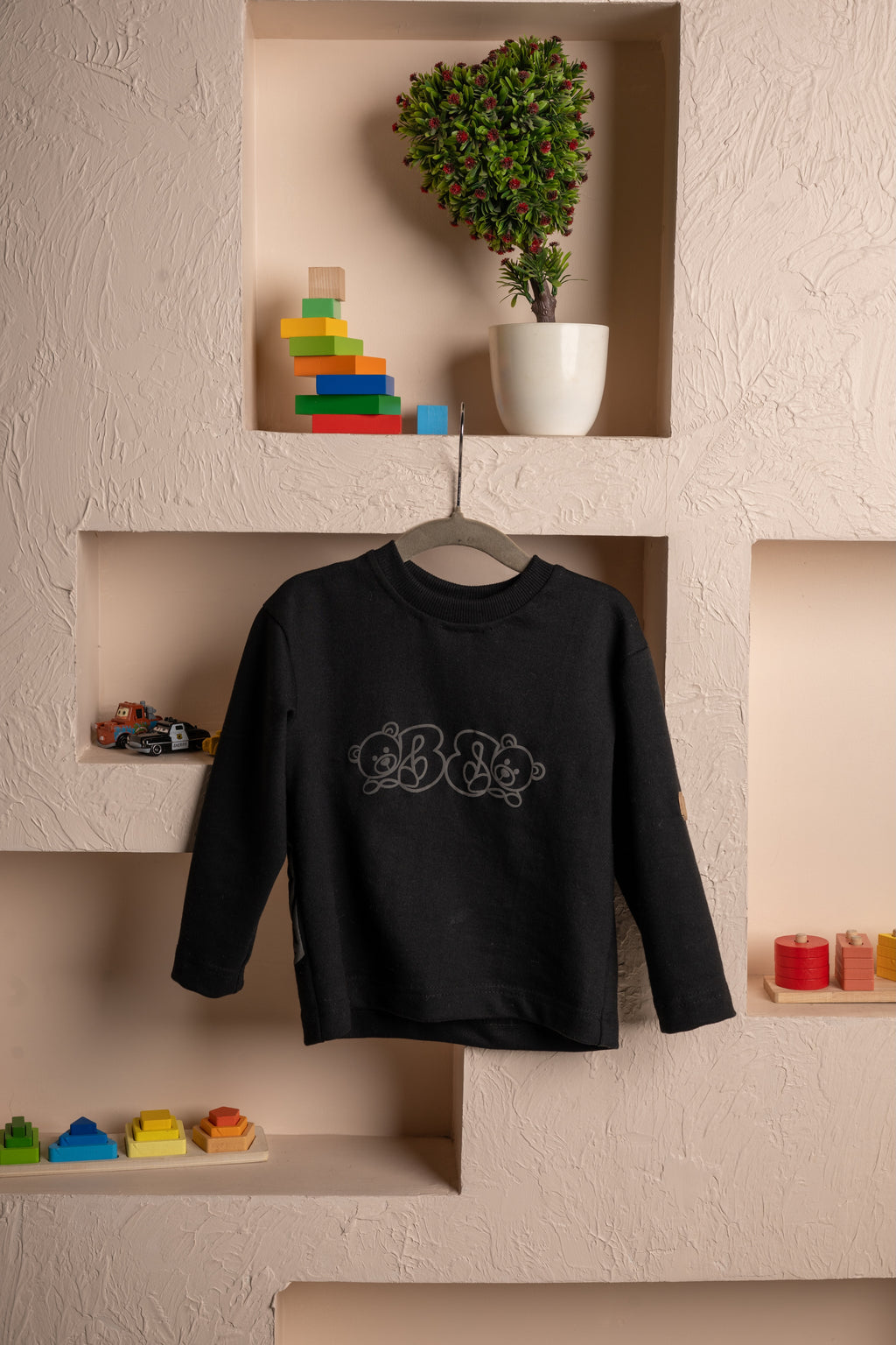 Elegant black children's sweater with subtle bear design displayed against textured beige wall with colorful wooden toys and potted plant